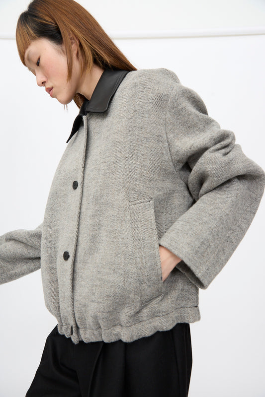 A person wearing a grey herringbone Studio Nicholson Seberg Chore Jacket in Black and White with a leather collar and black buttons, hands in the front pockets.