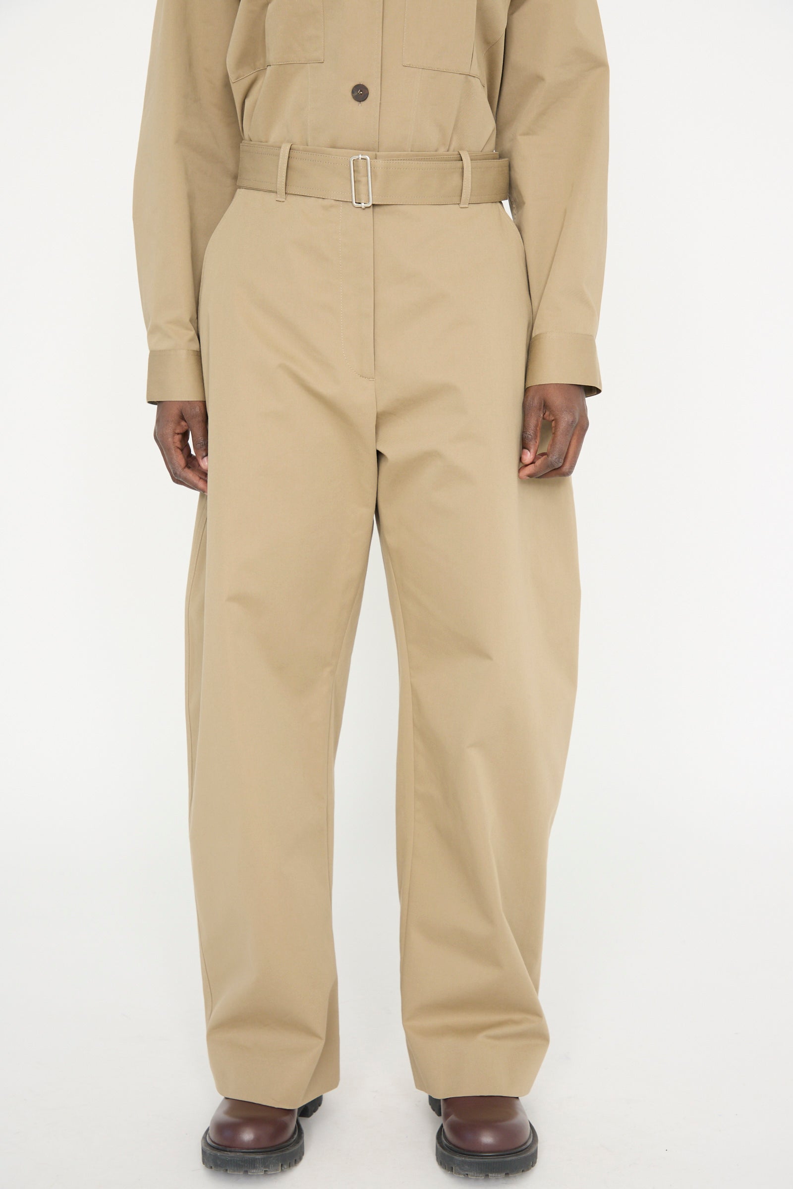 A person stands against a plain white background, wearing a beige button-up shirt and Studio Nicholson's Soft Technical Twill Doring Pant in Parcel, which are high-waisted and wide-legged with a belt.