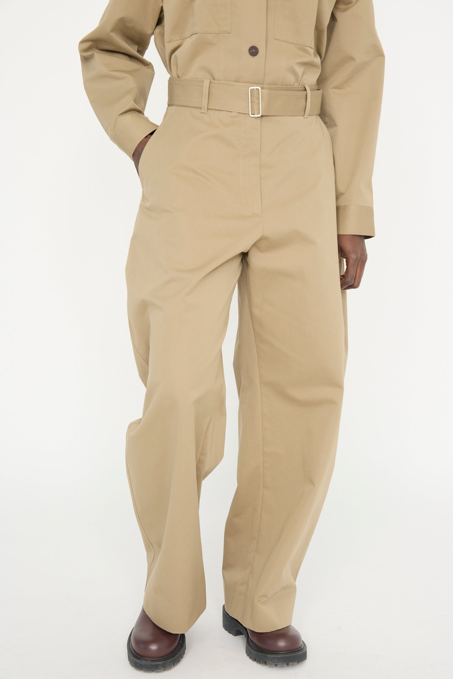 Against a plain background, a model wears Studio Nicholson's Soft Technical Twill Doring Pant in Parcel, featuring high-waist wide-leg pants and a belt, with one hand in a pocket. 