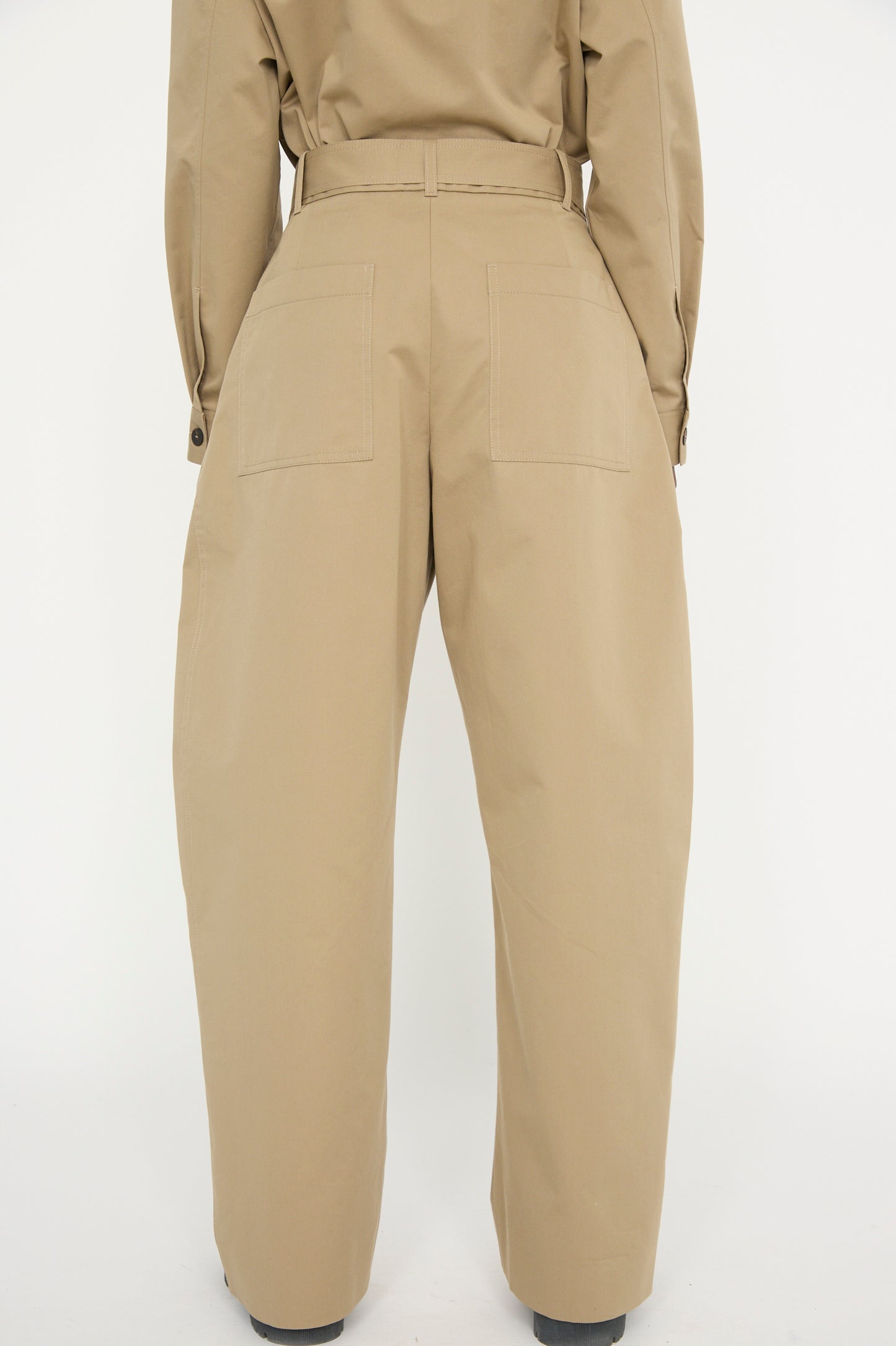 Back view of individual in Studio Nicholson's Soft Technical Twill Doring Pant in Parcel, featuring beige high-waisted, wide-leg design with patch pockets, set against a plain background.