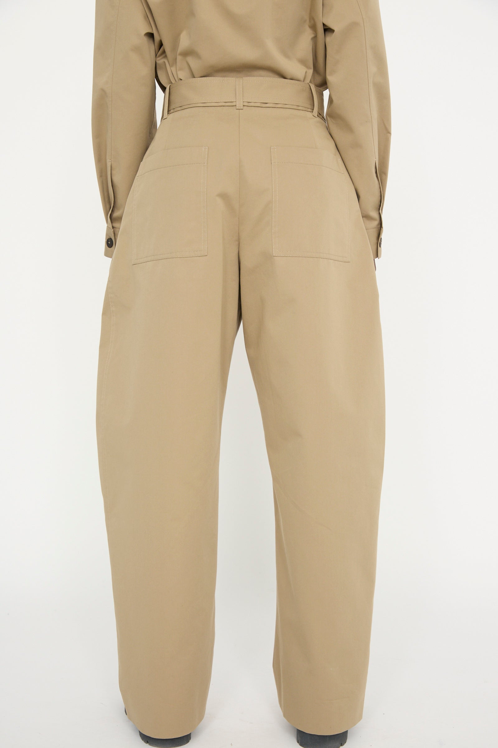 Back view of individual in Studio Nicholson's Soft Technical Twill Doring Pant in Parcel, featuring beige high-waisted, wide-leg design with patch pockets, set against a plain background.