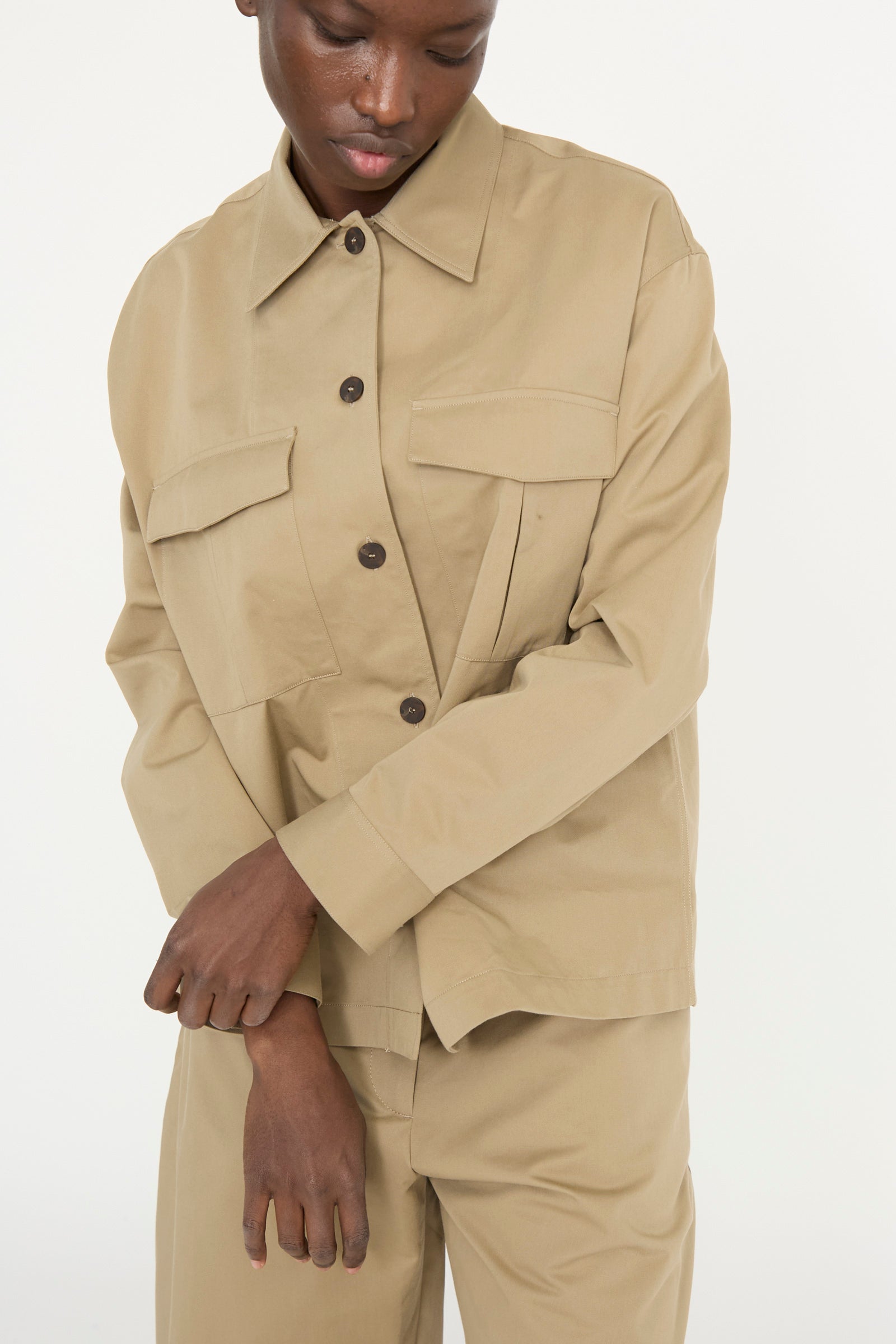 A person wearing the Studio Nicholson Soft Technical Twill Kukes Over Shirt in Parcel, featuring a beige military-style design with buttoned pockets and a boxy fit, gazes downward against a plain background.