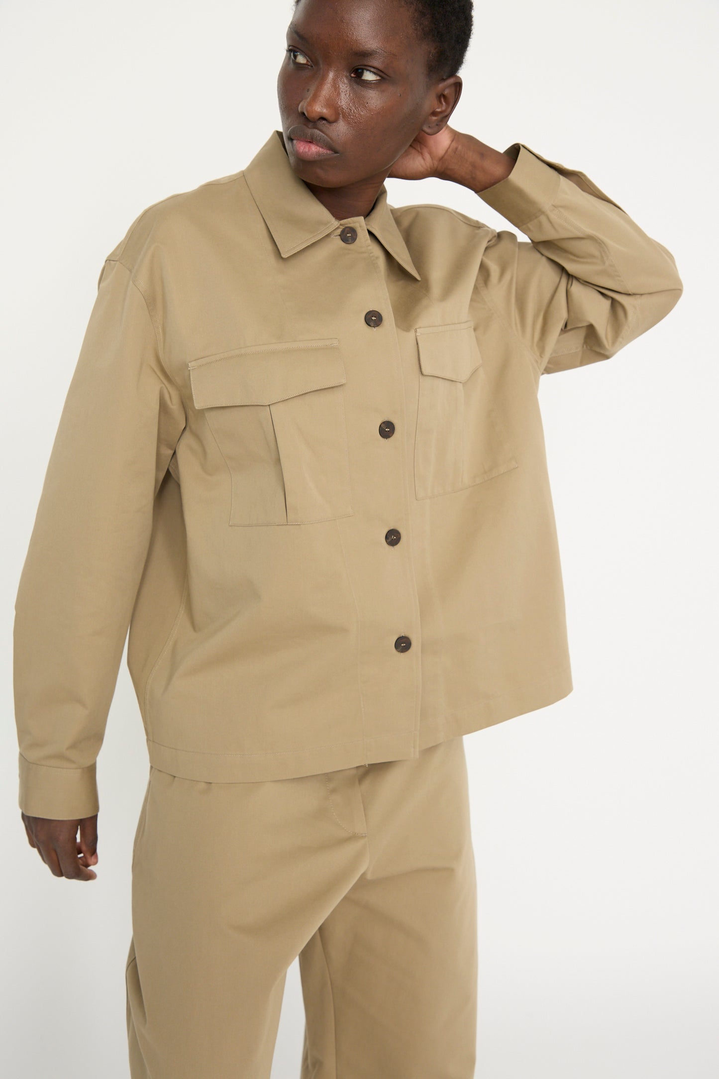 A person wearing the Studio Nicholson Soft Technical Twill Kukes Over Shirt in Parcel, with matching beige pants, poses against a plain background with one arm raised. 