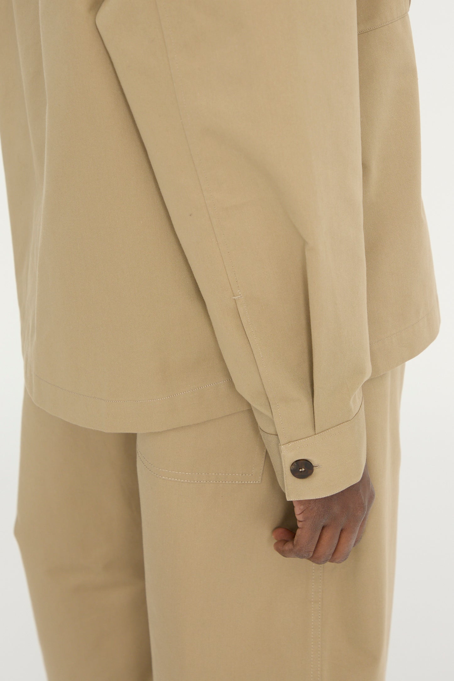 Close-up of someone wearing the Studio Nicholson Soft Technical Twill Kukes Over Shirt in Parcel, featuring a military style and buttoned cuff. Only the torso, arm, hand, and part of their pants are visible.