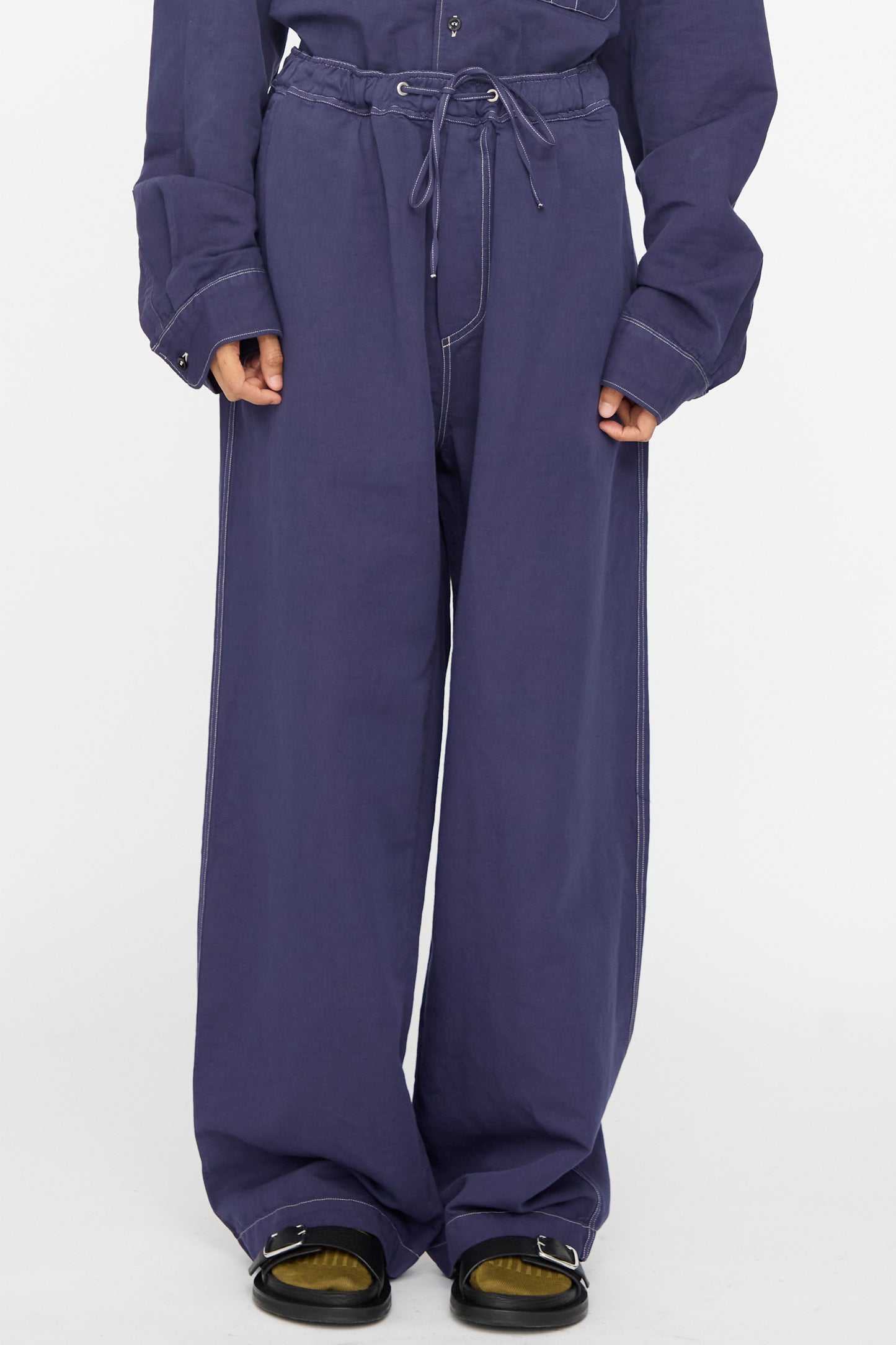 A person wears the Studio Nicholson Sonny Drawstring Pant in Indigo with a matching long-sleeve top and black sandals, standing against a plain white background.