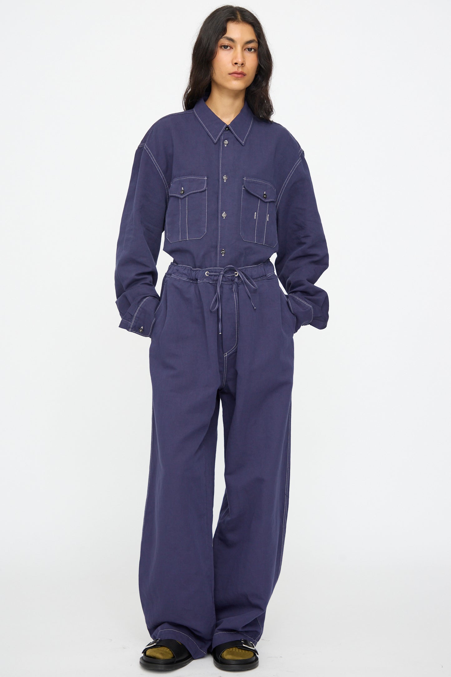 A person stands facing forward against a plain white background, wearing Studio Nicholson's Sonny Drawstring Pant in Indigo paired with black shoes. The pants are loose-fitting, long, and feature an elastic waistband.