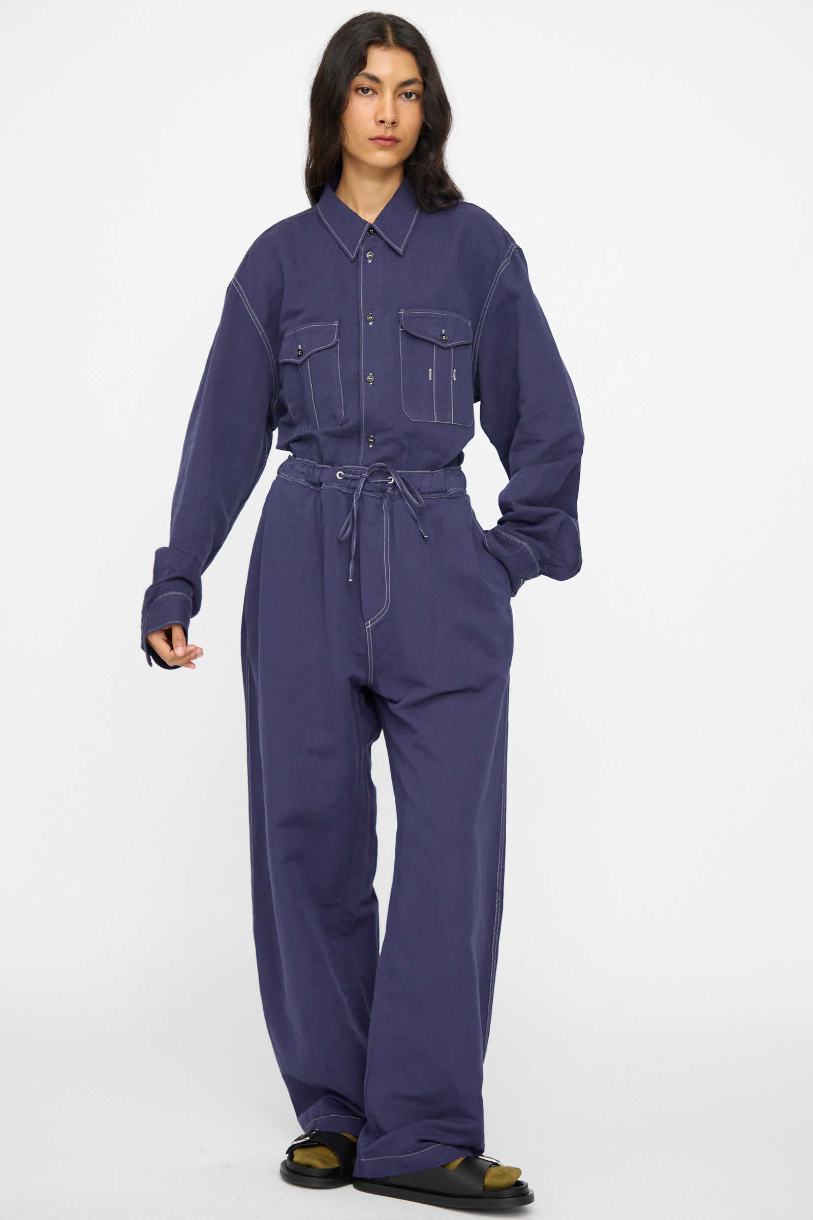 A person stands facing forward wearing Studio Nicholson’s Sonny Drawstring Pant in Indigo, styled with black sandals.