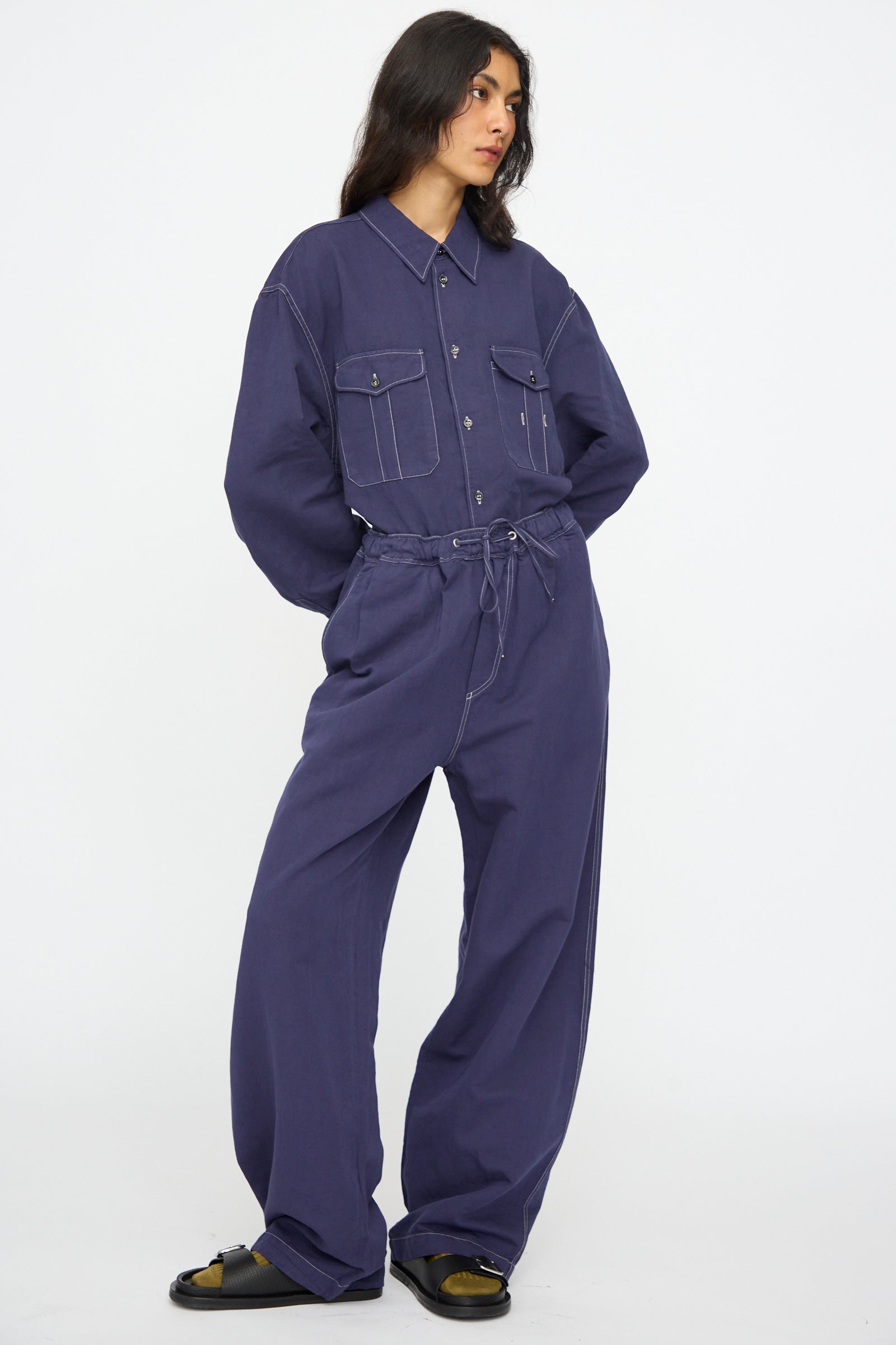 A person wears a loose-fitting, dark blue button-up shirt and Studio Nicholson Sonny Drawstring Pant in Indigo with an elastic waistband, posing against a plain white background.