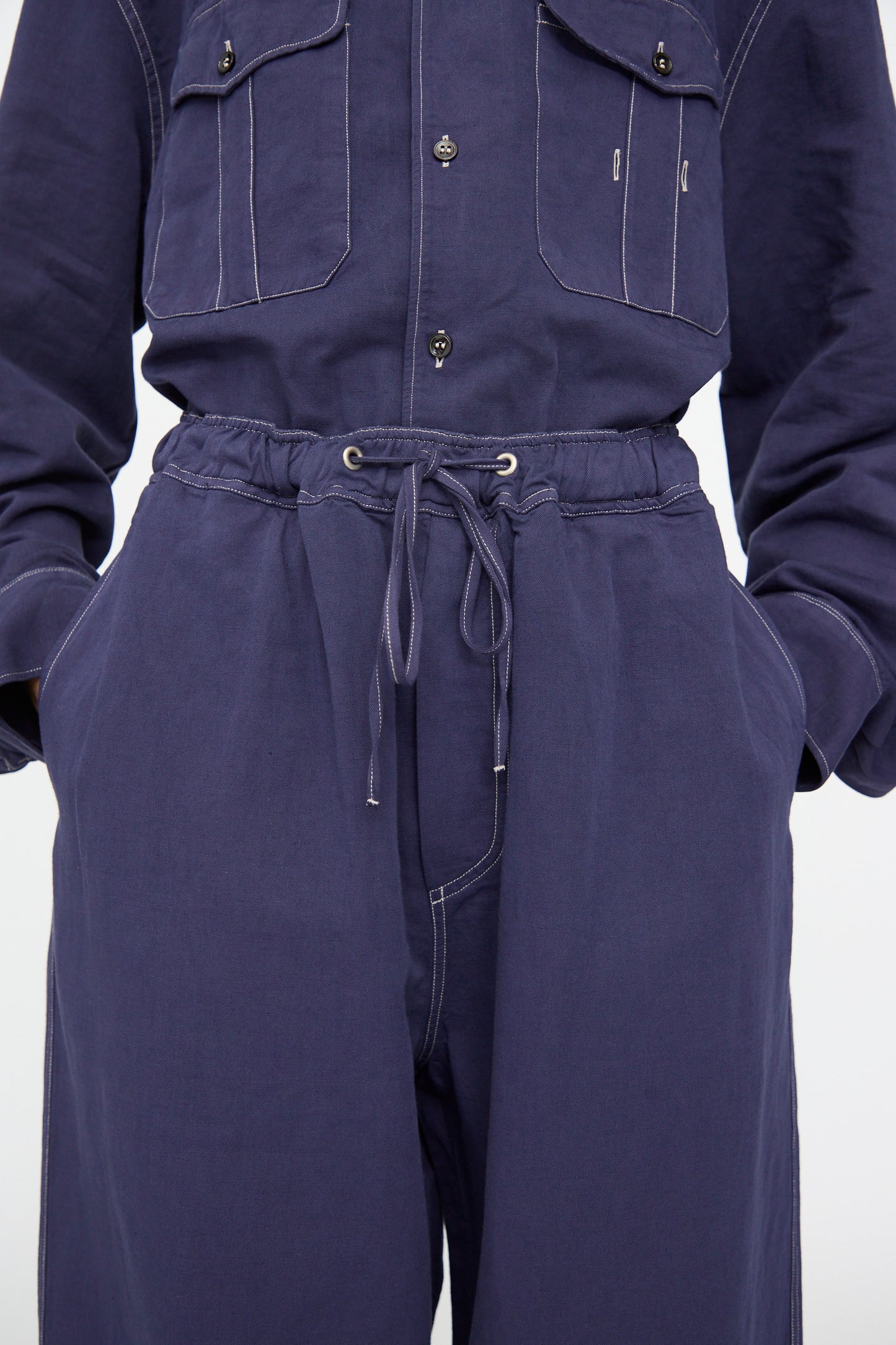 Model wears the Studio Nicholson Sonny Drawstring Pant in Indigo, featuring wide legs, a drawstring waist, white stitching, and buttoned pockets. Paired with hands in pockets for a relaxed look.