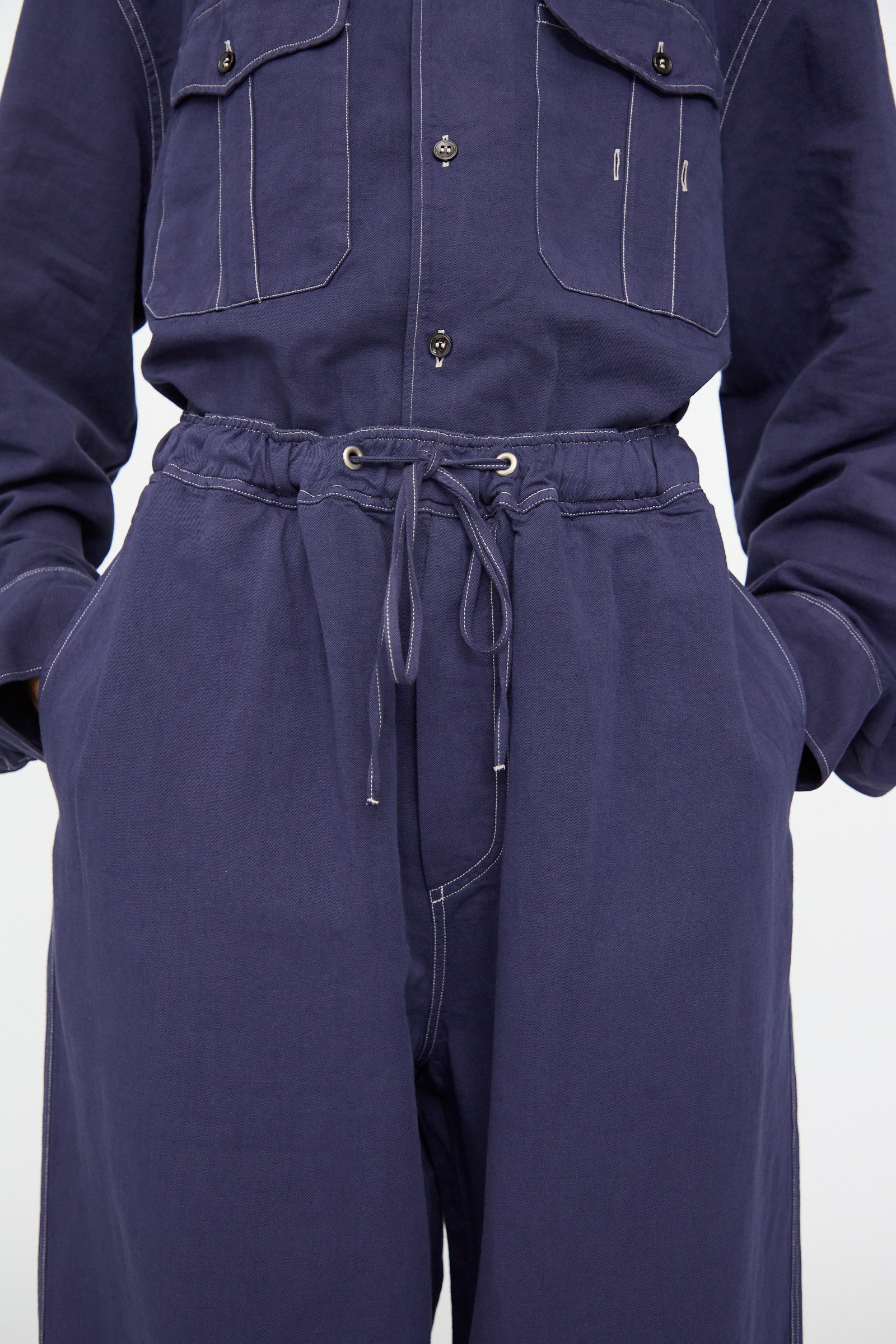 Model wears the Studio Nicholson Sonny Drawstring Pant in Indigo, featuring wide legs, a drawstring waist, white stitching, and buttoned pockets. Paired with hands in pockets for a relaxed look.