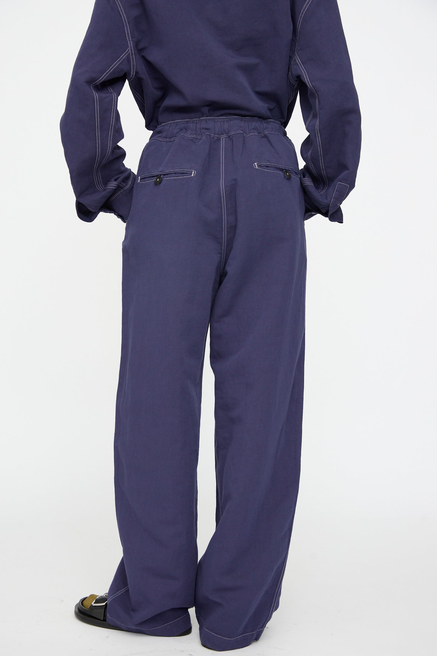Person wearing Studio Nicholson’s Sonny Drawstring Pant in Indigo, a loose-fitting linen style with an elastic waistband, matched with a coordinating top, seen from the back against a plain white background.