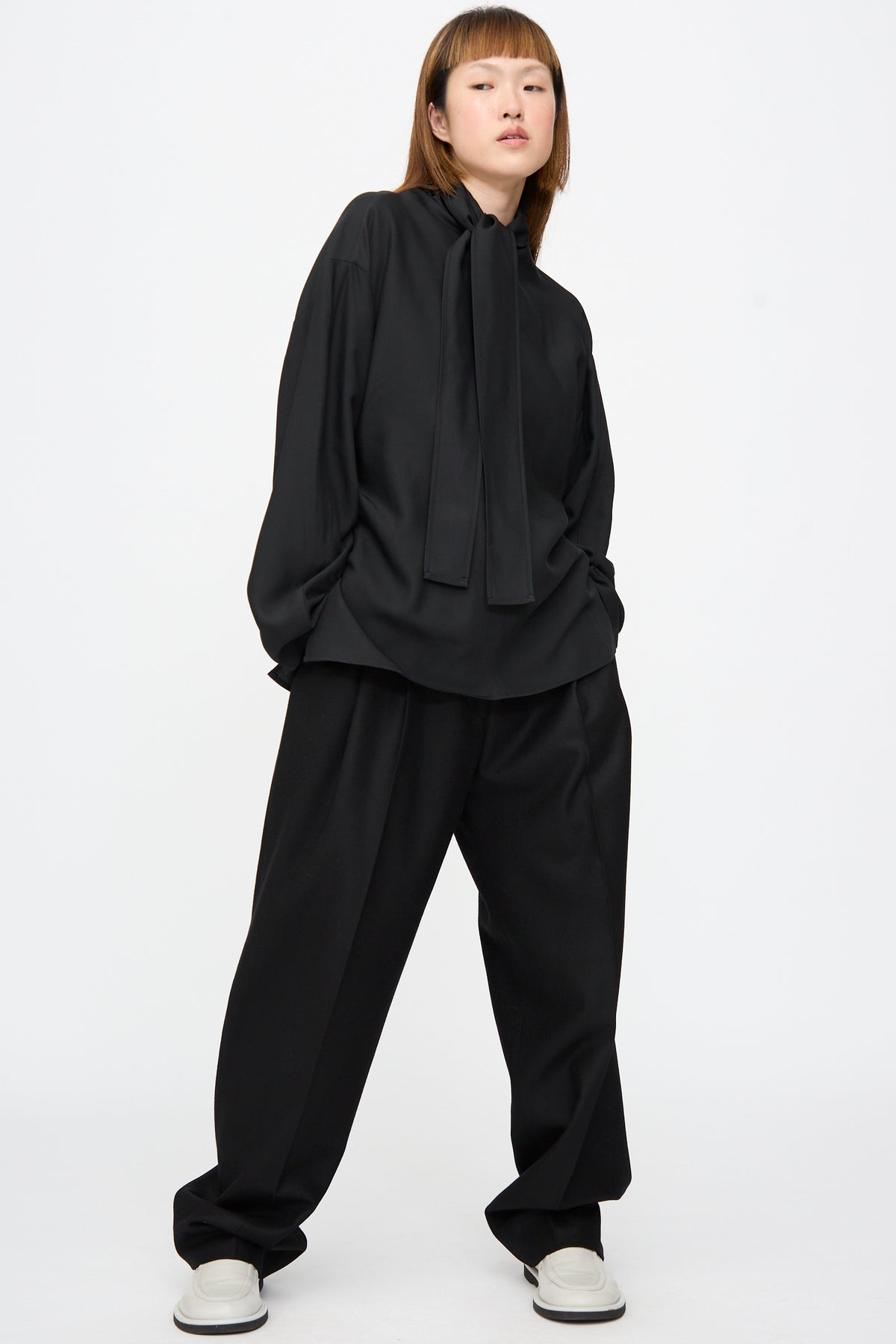 A person wears a black long-sleeved shirt with a neck tie and Studio Nicholson Sperro Double Pleat Pant in black.