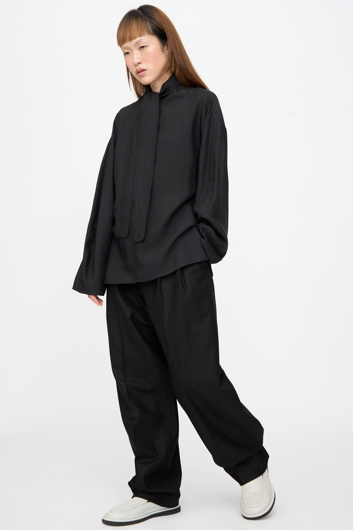 A person wearing a black long-sleeved mock-neck shirt and black Sperro Double Pleat Pant by Studio Nicholson, paired with white loafers.