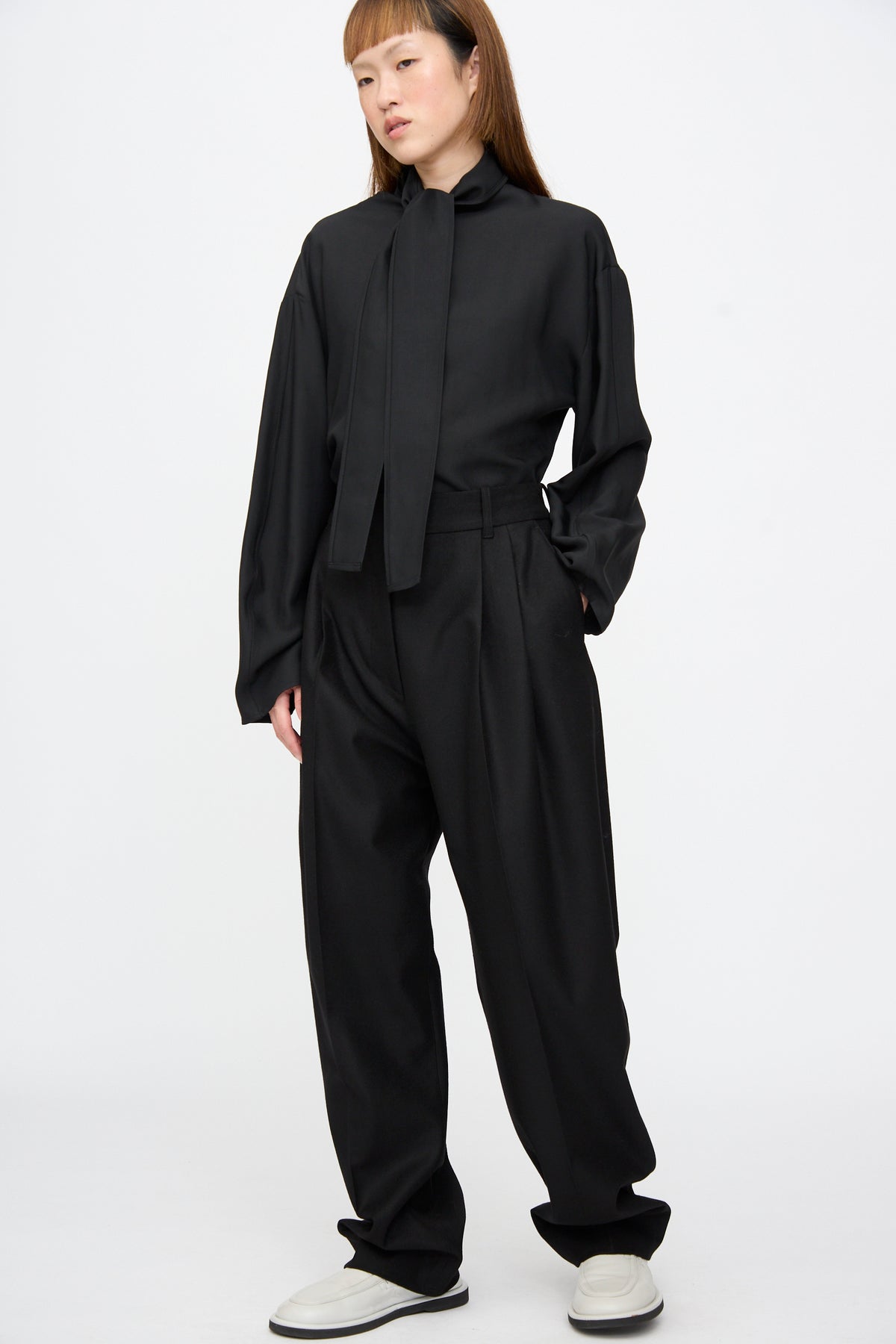 A person wearing a black long-sleeved blouse with a neck tie, Studio Nicholson Sperro Double Pleat Pant in Black, and white shoes.