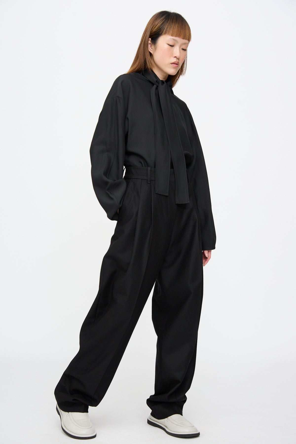 A person wearing a black long-sleeved tie-neck blouse and Studio Nicholson Sperro Double Pleat Pant in Black, featuring a high-waisted, double-pleated, tapered leg.