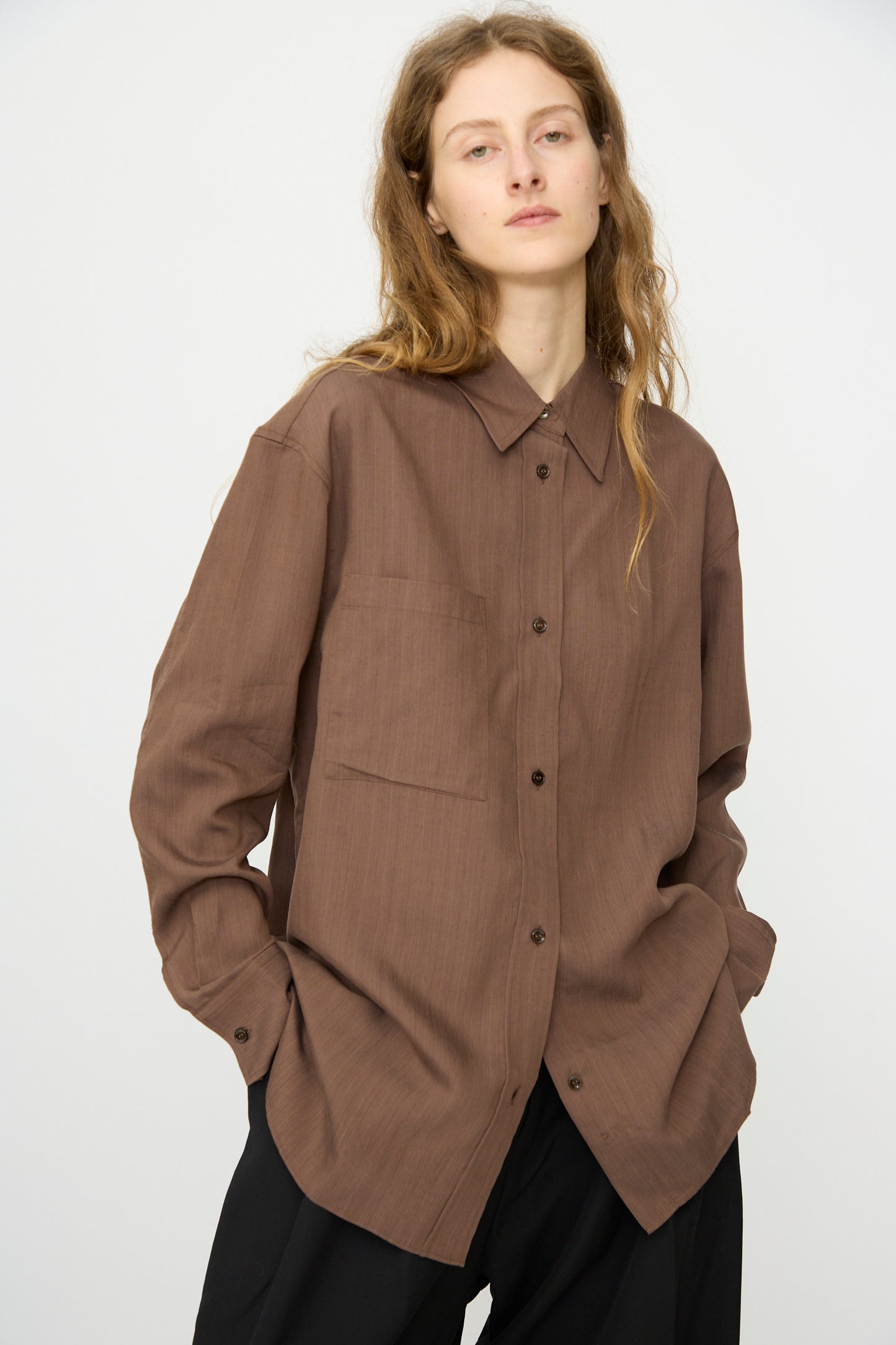 A person with long, wavy hair wears the Subtle Pinstripe Leto Shirt in Brown by Studio Nicholson and black pants, standing against a plain light background.