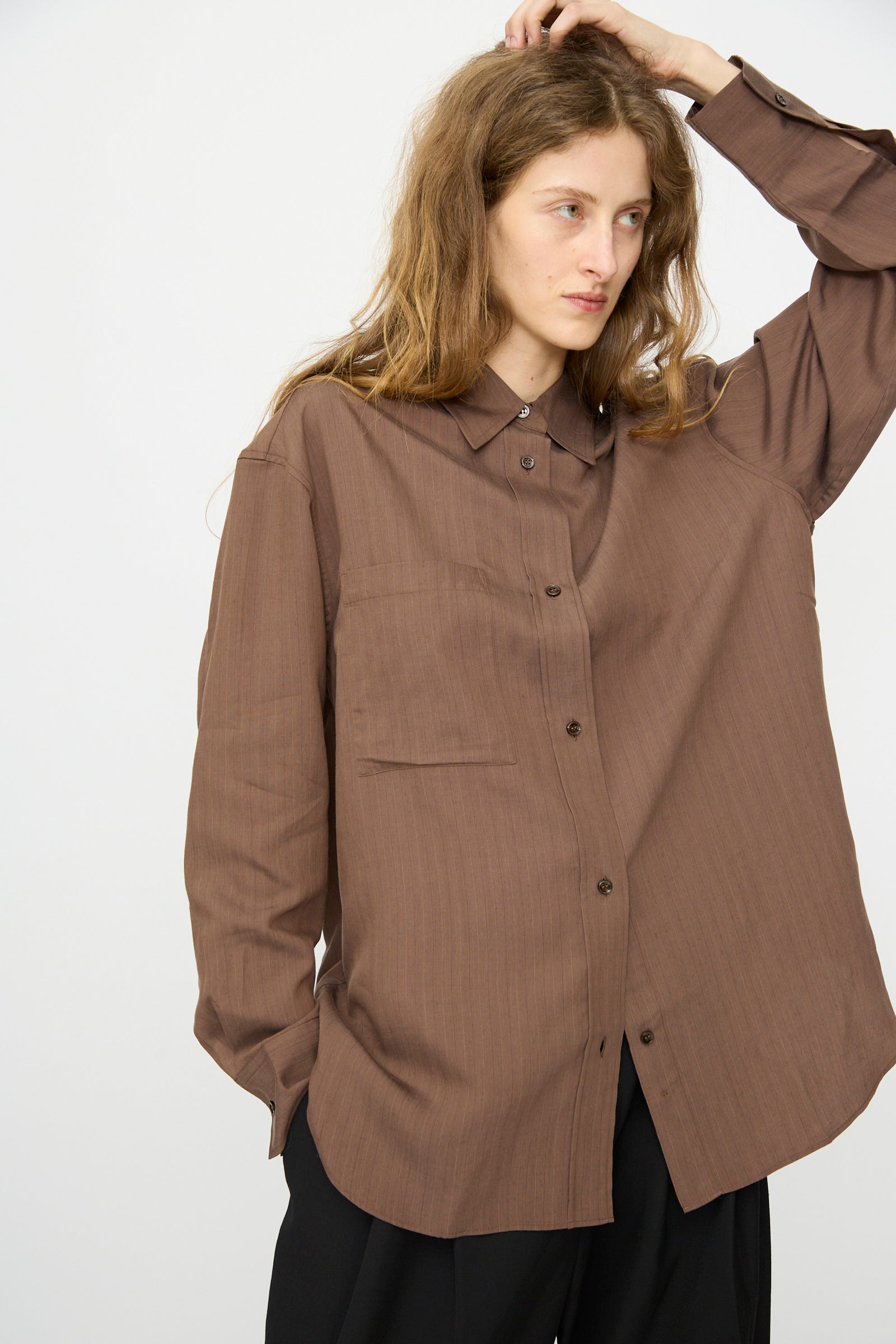 Person with long, wavy hair wears Studio Nicholson's Subtle Pinstripe Leto Shirt in Brown and black pants, standing against a plain light background with one hand raised to their head.