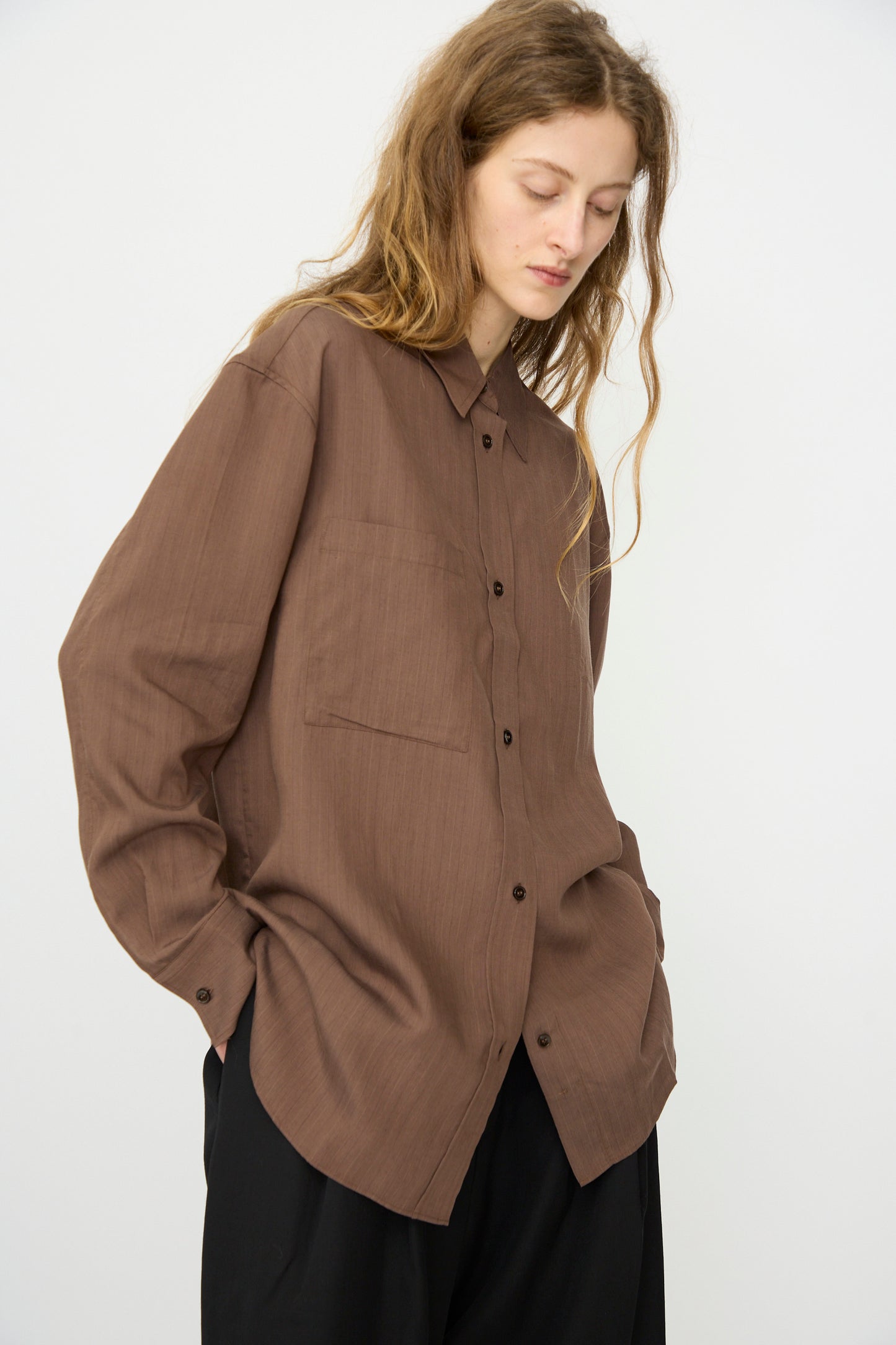 A person with long, wavy hair wears the Subtle Pinstripe Leto Shirt in Brown by Studio Nicholson with black pants, standing against a plain white background.