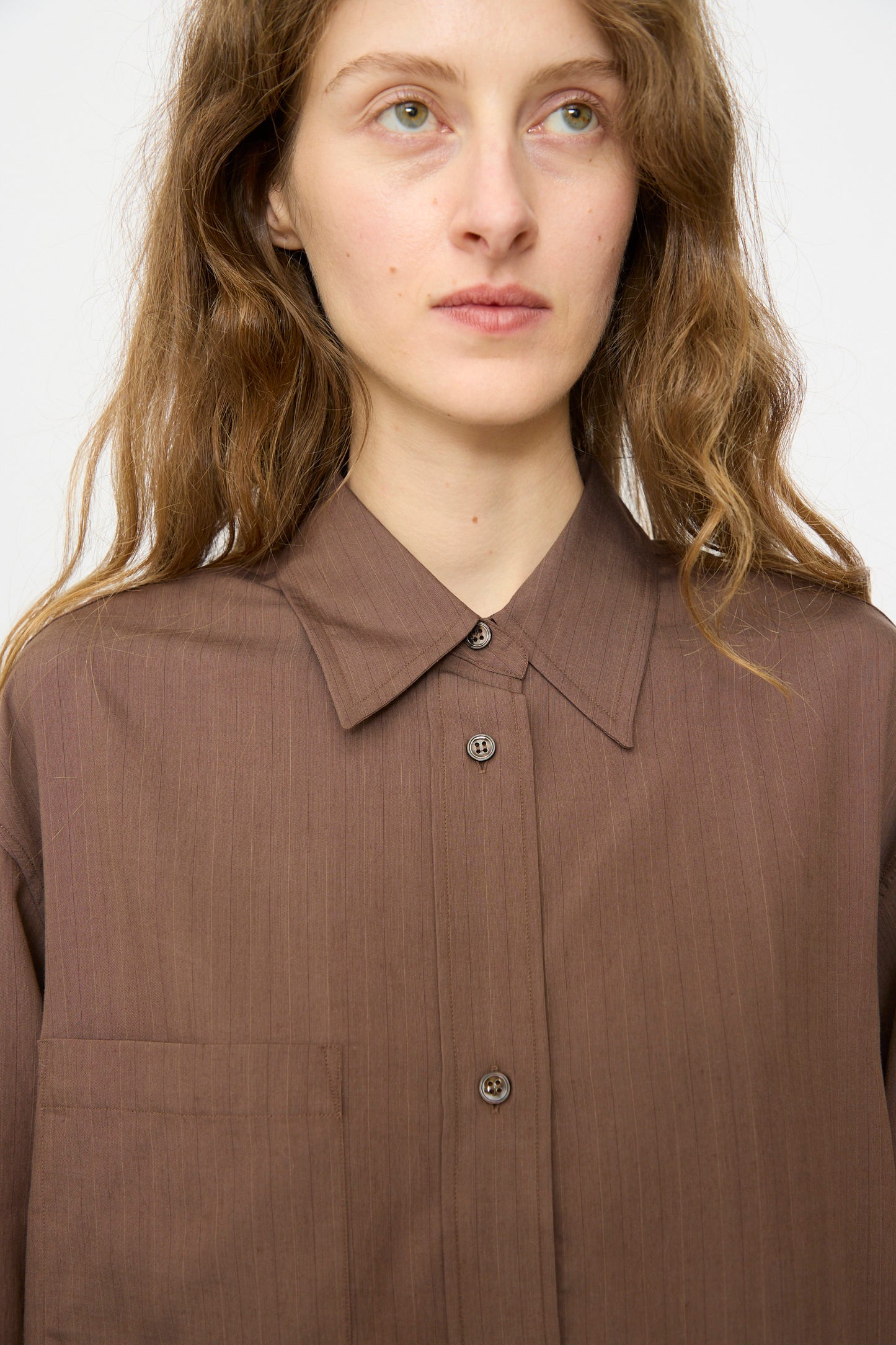 A person with wavy brown hair wears the Studio Nicholson Subtle Pinstripe Leto Shirt in Brown—an oversized button-up with a chest pocket—standing against a plain light background.
