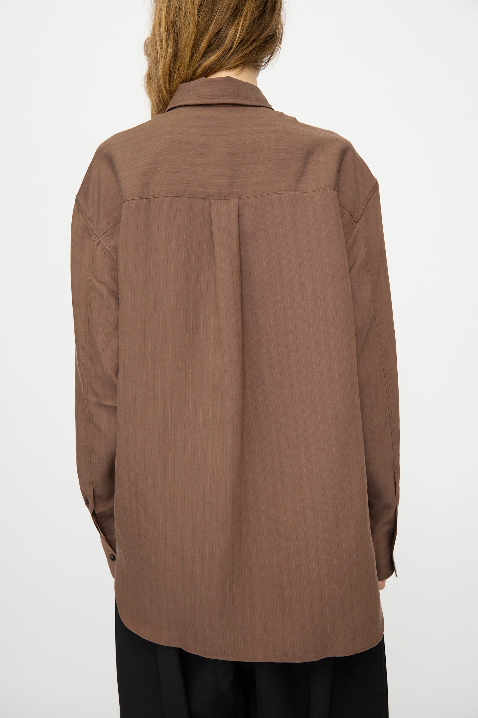 A person stands with their back to the camera, wearing the Studio Nicholson Subtle Pinstripe Leto Shirt in brown and black pants against a plain white background.