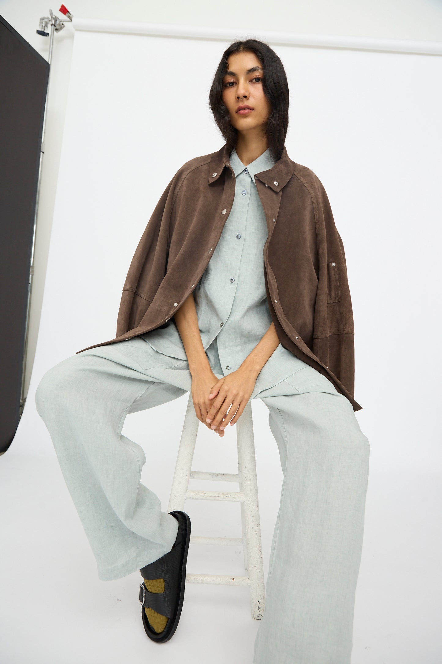 A person sits on a white stool against a plain white background, wearing a light gray outfit with the Studio Nicholson Suede Utility Dash Jacket in Grey draped over their shoulders.