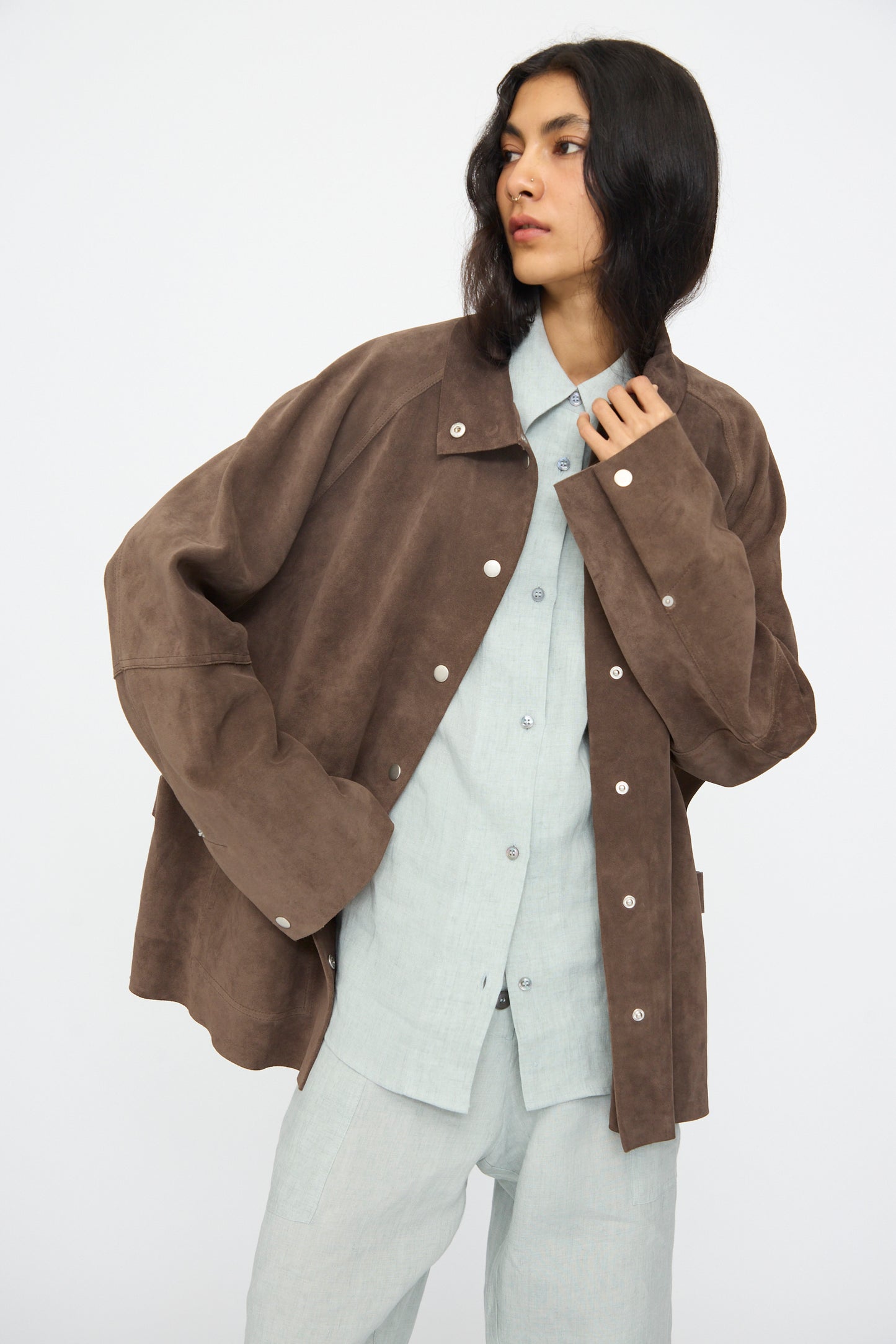 Model wears the Studio Nicholson Suede Utility Dash Jacket in Grey over a light gray button-up shirt and pants, standing against a plain white background.