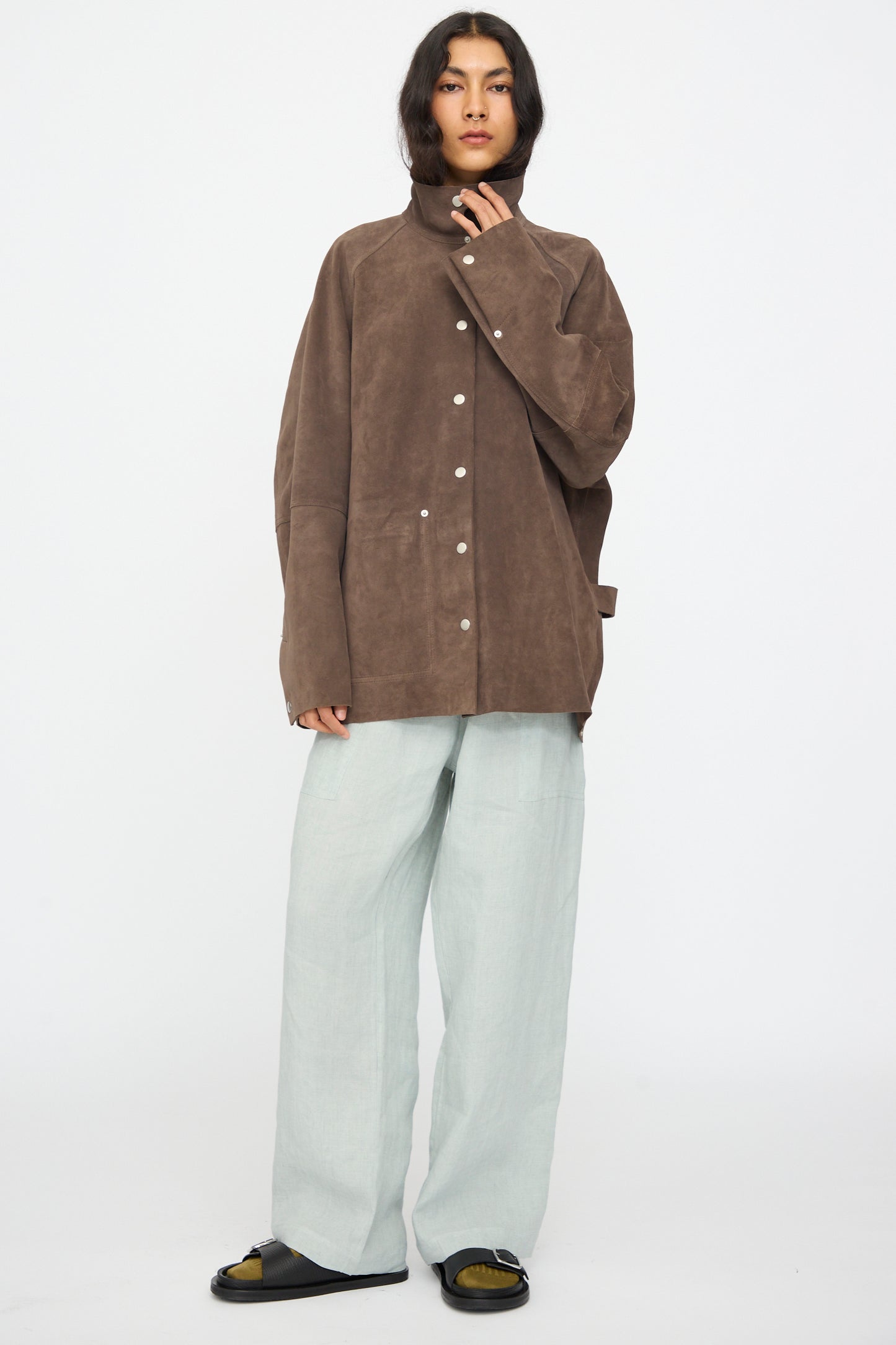 A person stands against a plain white background wearing the Studio Nicholson Suede Utility Dash Jacket in Grey, light gray wide-leg pants, and black slide sandals.