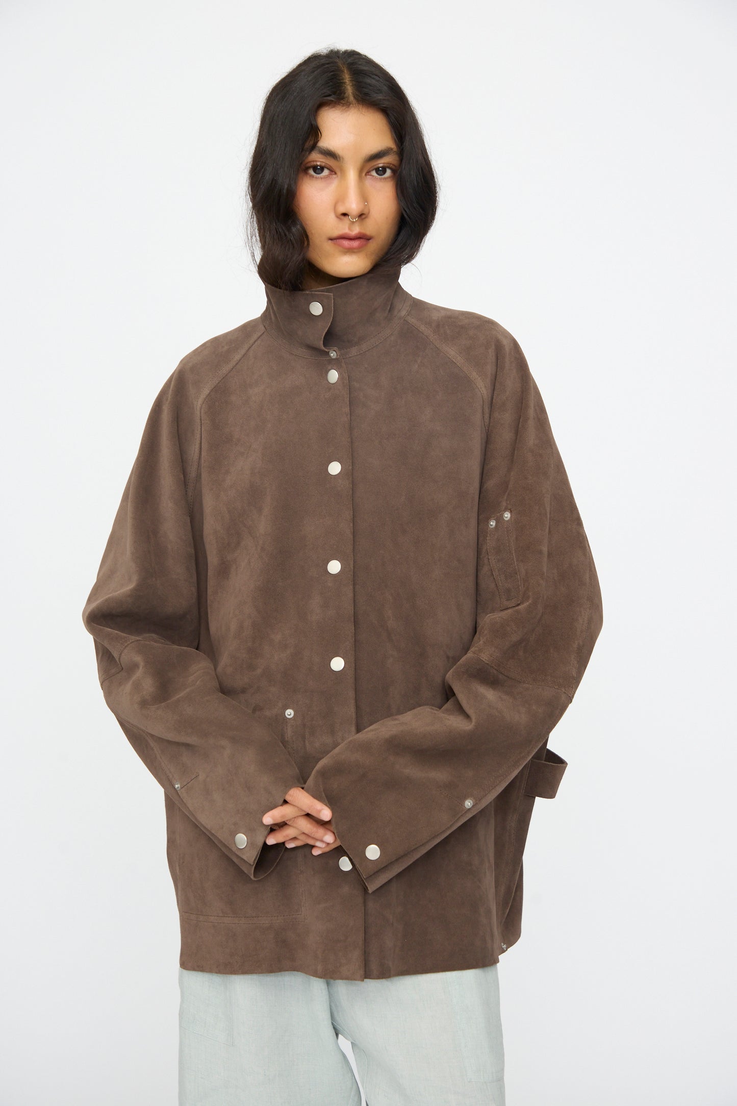A person stands facing forward against a plain white background, wearing Studio Nicholson's Suede Utility Dash Jacket in Grey and light-colored pants.