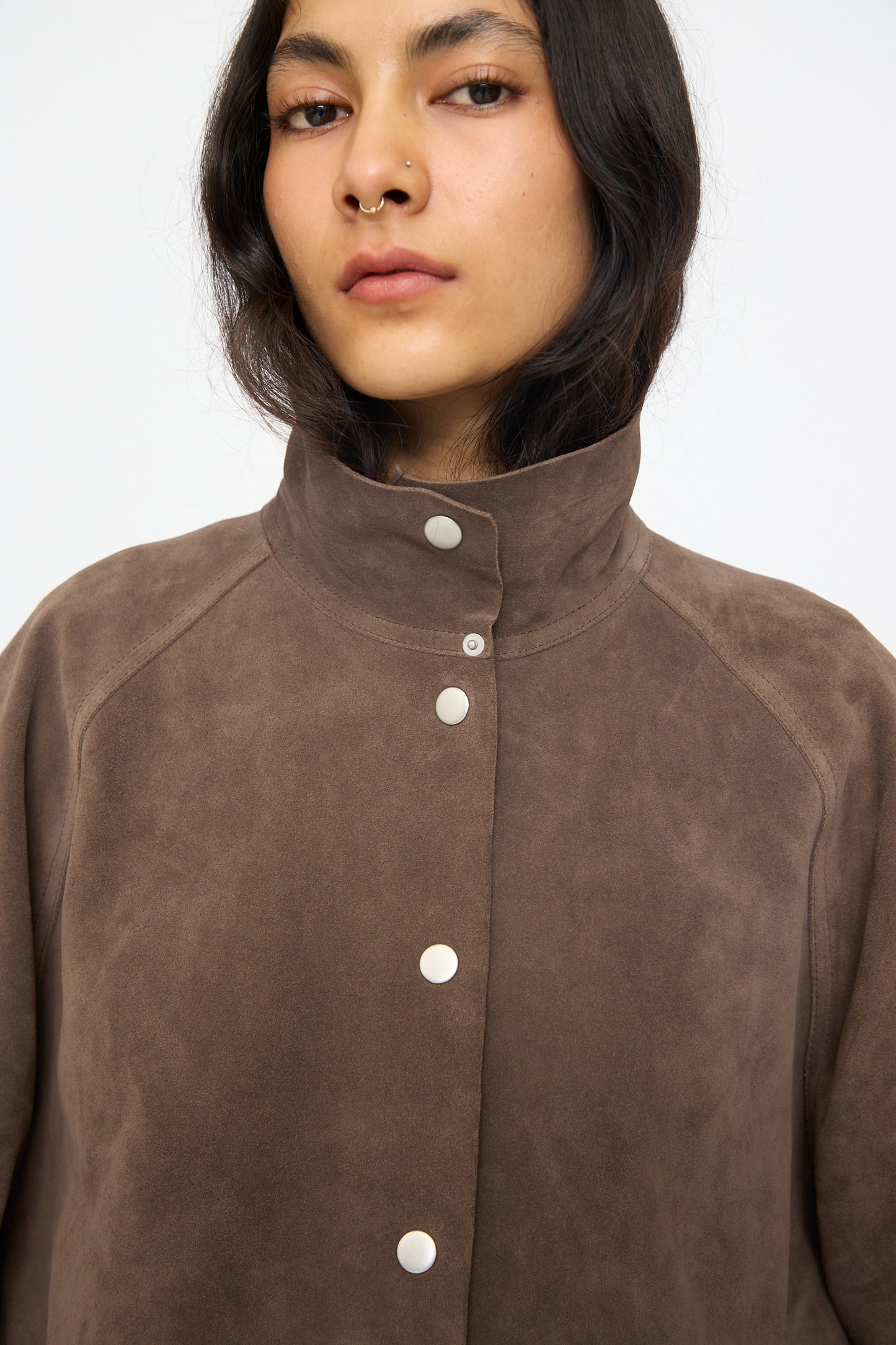 A person with long dark hair and a nose ring wears the Studio Nicholson Suede Utility Dash Jacket in Grey, featuring a high collar and silver snap buttons, gazing at the camera.