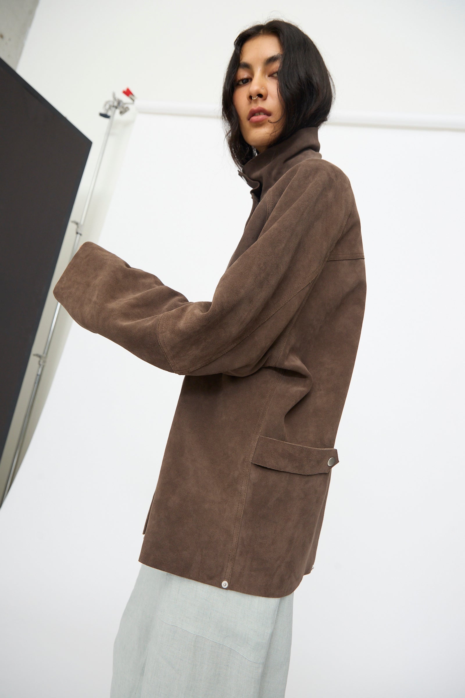 A woman in the Studio Nicholson Suede Utility Dash Jacket in Grey and light gray pants stands sideways against a white background, looking toward the camera.