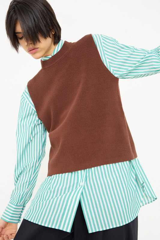 A person posing in a layered fashion outfit, featuring a Studio Nicholson Sumire Sleeveless Top in Carob Brown over a teal and white striped long-sleeve shirt, paired with black trousers.