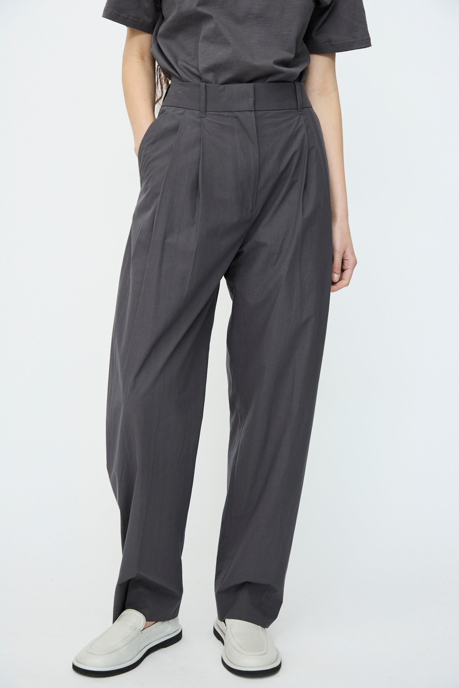 Dressed in Studio Nicholson's Thunder Typewriter Cotton Sperro Pant and a T-shirt, a person stands against a plain white background, paired with white slip-on shoes.