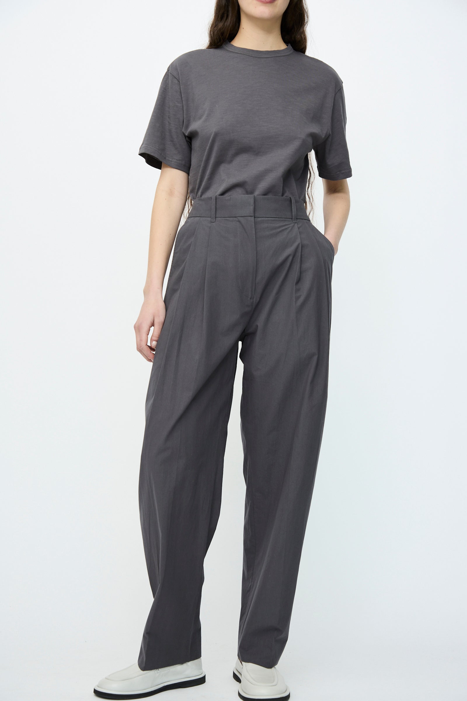 In Studio Nicholson, an individual wears Thunder Typewriter Cotton Sperro Pants, high-waisted and relaxed fit, along with a gray T-shirt and white shoes against a plain backdrop.