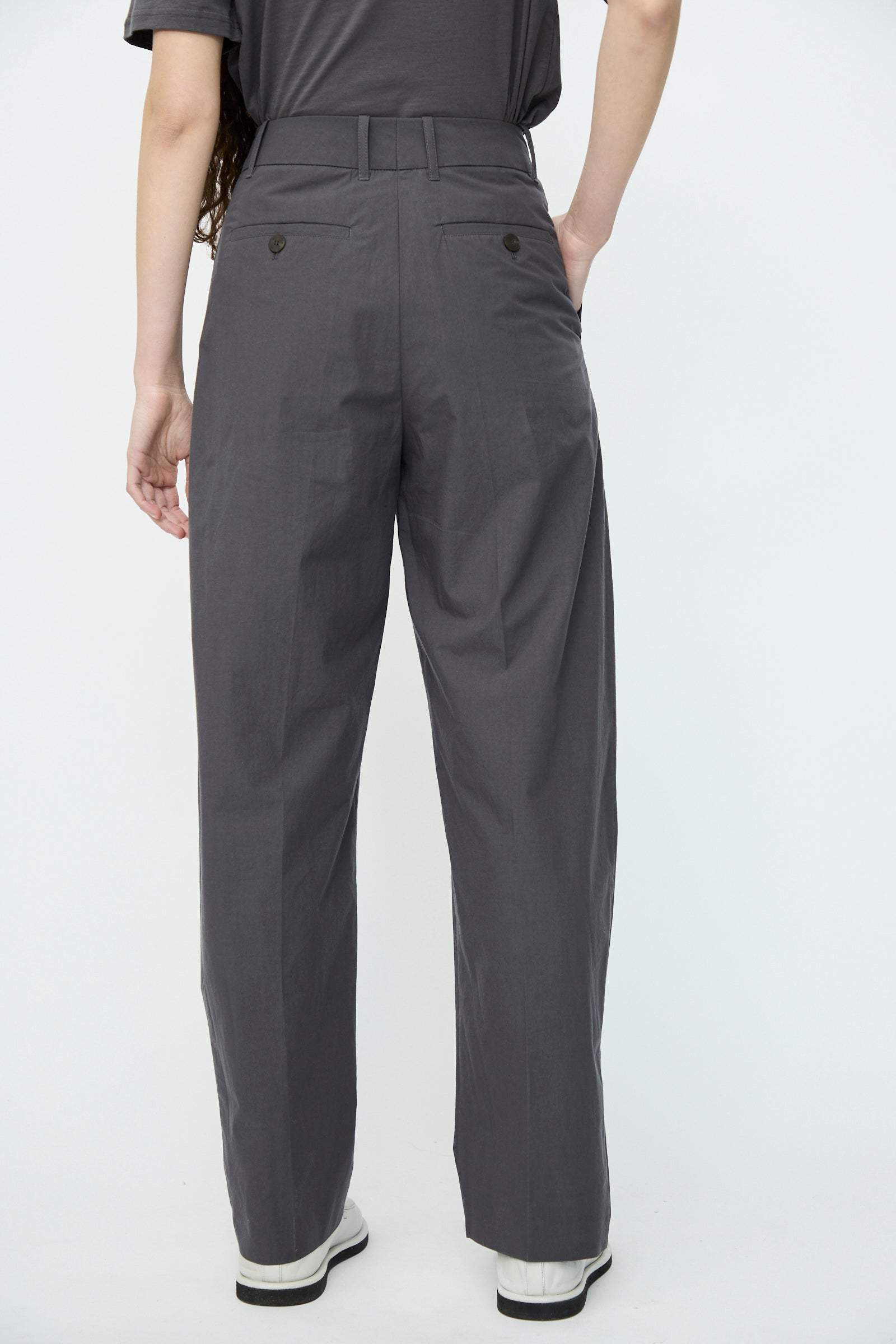 A person in Studio Nicholson's Typewriter Cotton Sperro Pant in Thunder and white sneakers stands with one hand in the pocket, viewed from the back.