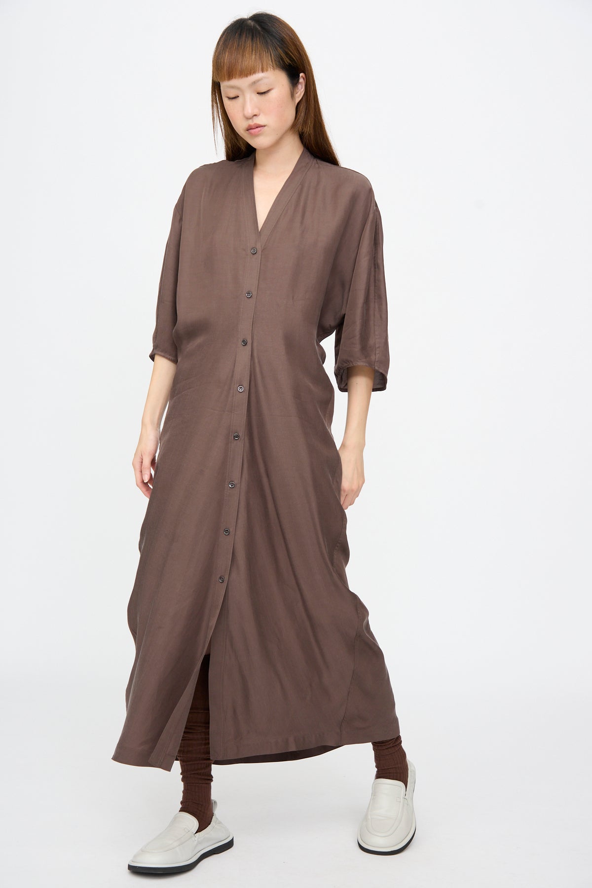 A person models the Studio Nicholson Yosha Dress in Porchini, a long, brown, button-front V-neck maxi dress with elbow-length sleeves, worn with brown leggings and off-white loafers.