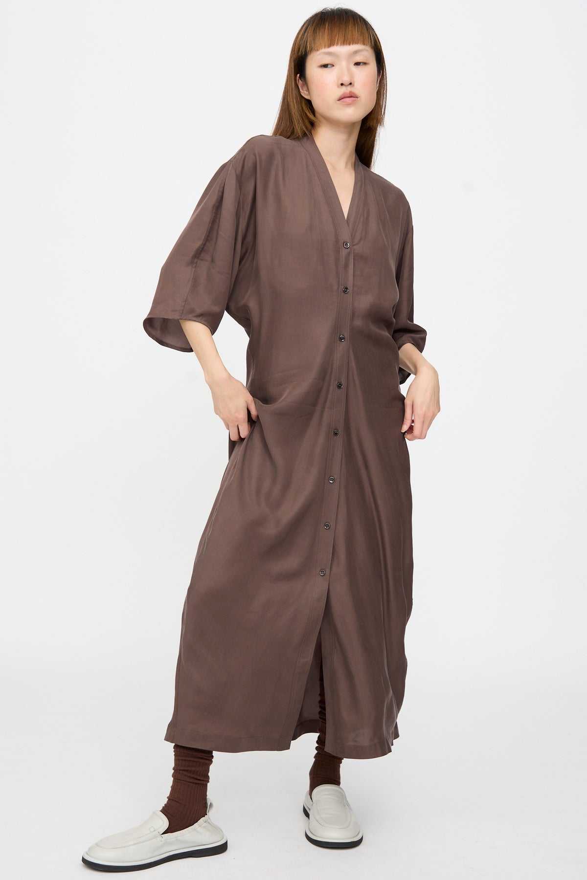 A model wears a brown Studio Nicholson Yosha Dress in Porchini with three-quarter sleeves, brown socks, and white loafers.