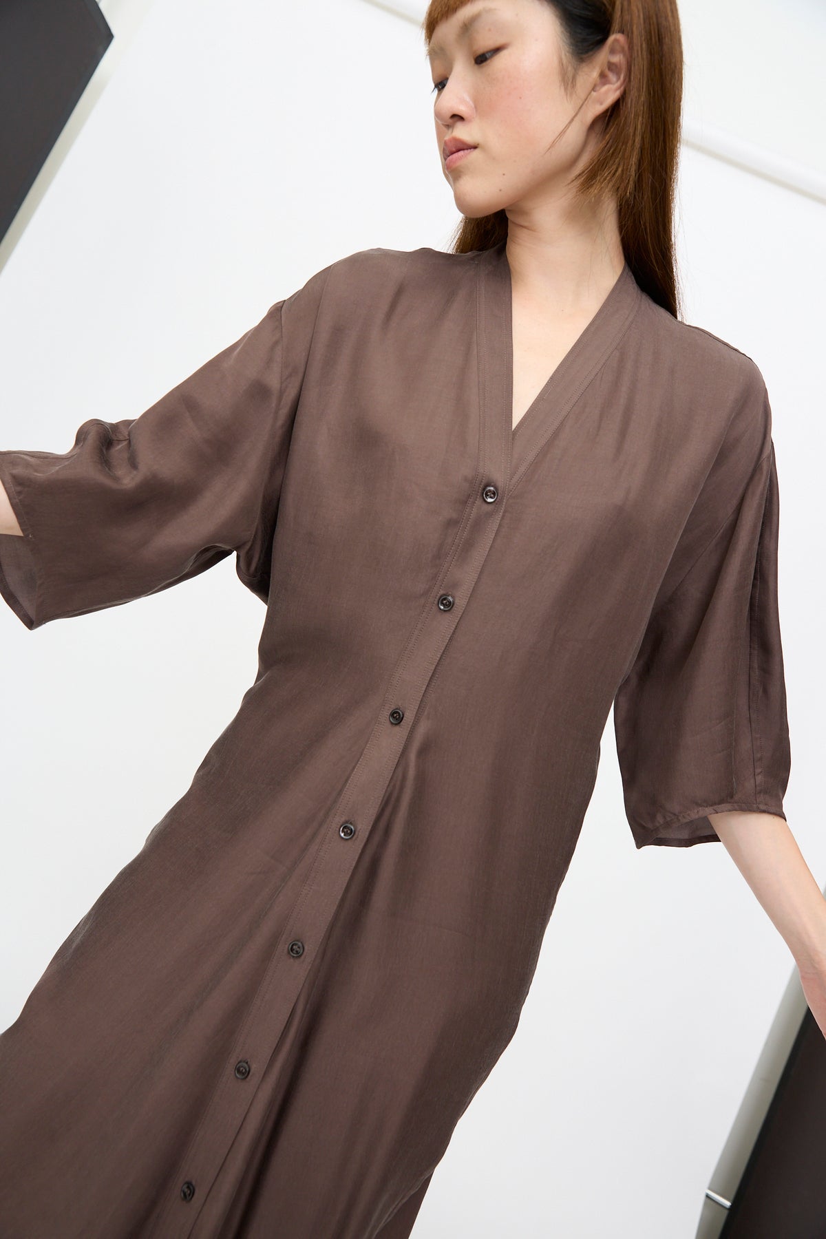 A person wearing a brown Studio Nicholson Yosha Dress in Porchini, an FSC lyocell button-down midi dress with a V-neck and elbow-length sleeves.