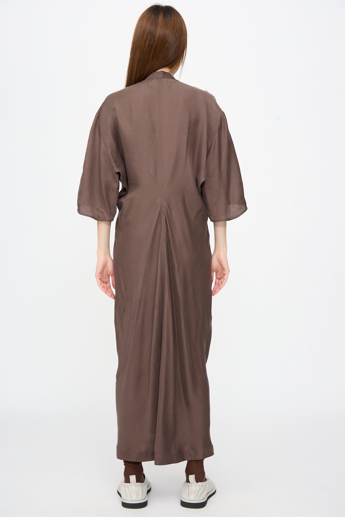 A person seen from the back wearing a dark brown, calf-length Studio Nicholson Yosha Dress in Porchini with elbow-length sleeves, brown socks, and white sneakers.