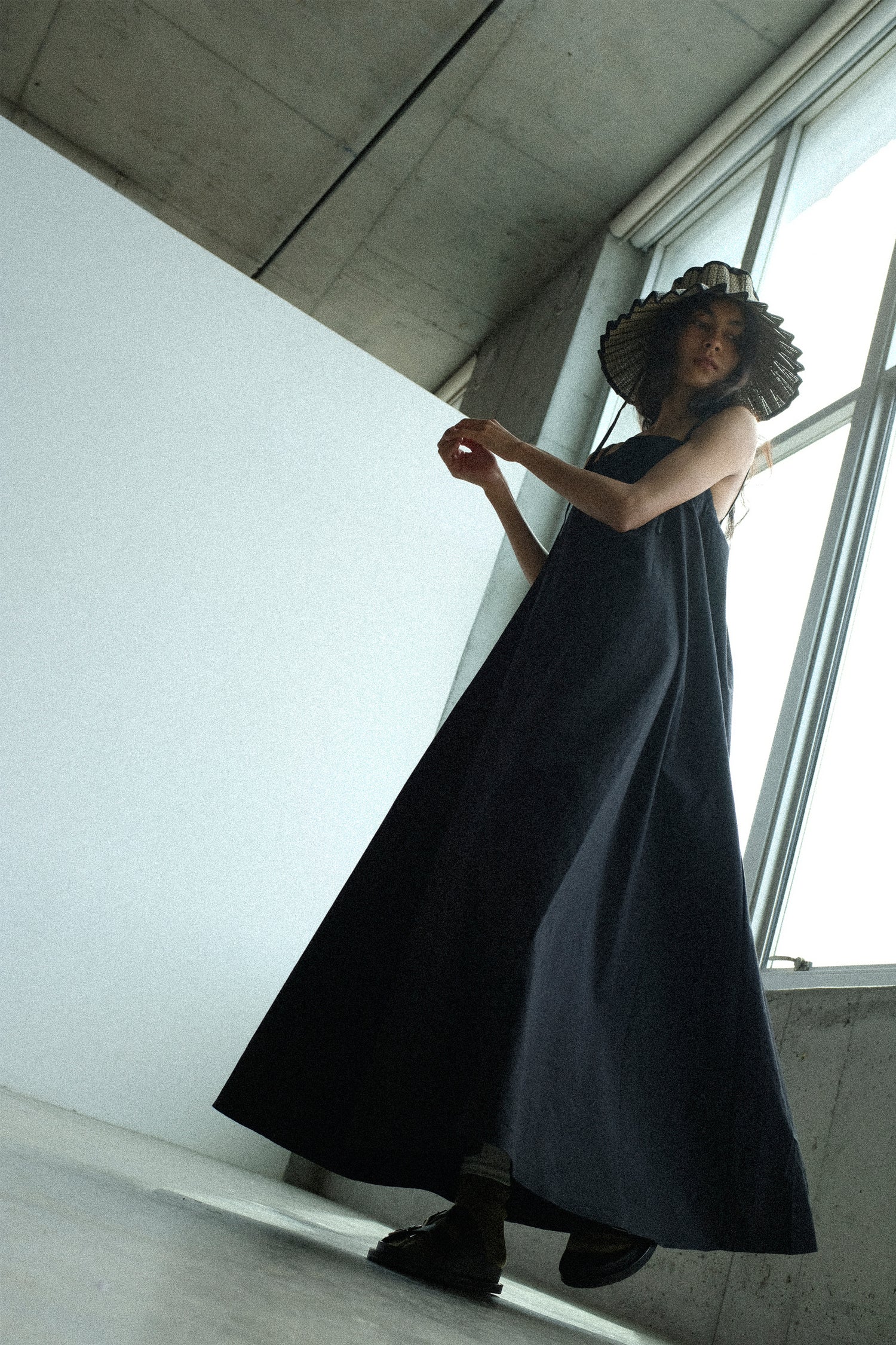 Woman in a long black dress standing indoors with large windows.
