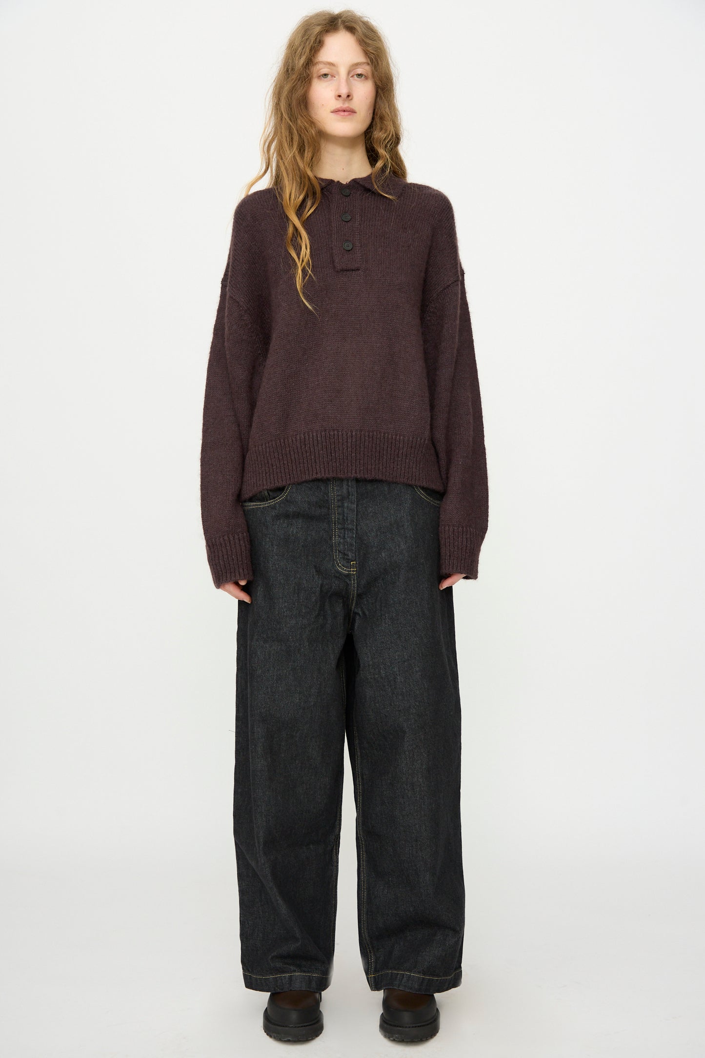 A person with long hair stands facing forward against a white background, wearing Studio Nicholson's Brushed Mohair Dune Knit Shirt in Espresso, loose dark jeans, and black shoes.