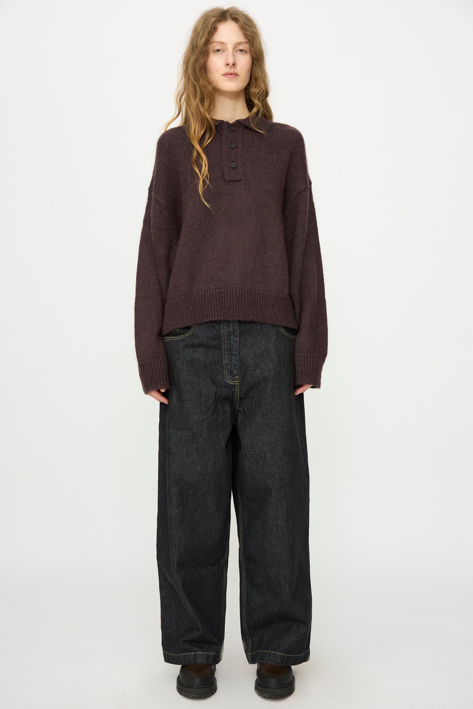 A person with long hair stands facing forward against a white background, wearing Studio Nicholson's Brushed Mohair Dune Knit Shirt in Espresso, loose dark jeans, and black shoes.