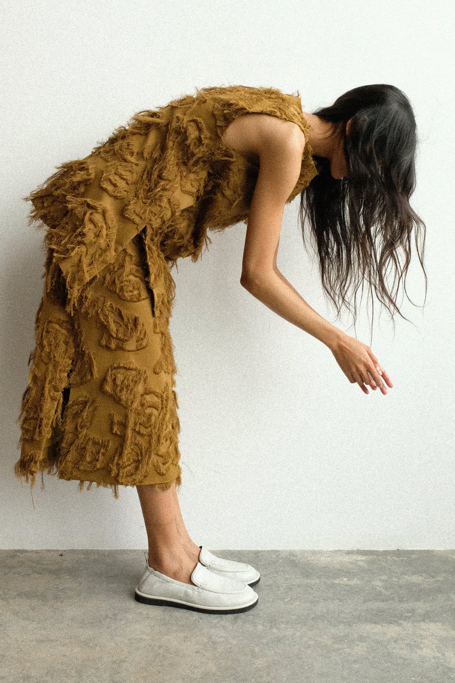 A woman reaching down wearing Studio Nicholson's Fringe Jacquard Mager Top in Mustard with the Fringe Jacquard Roeper Skirt in Mustard against a white wall.