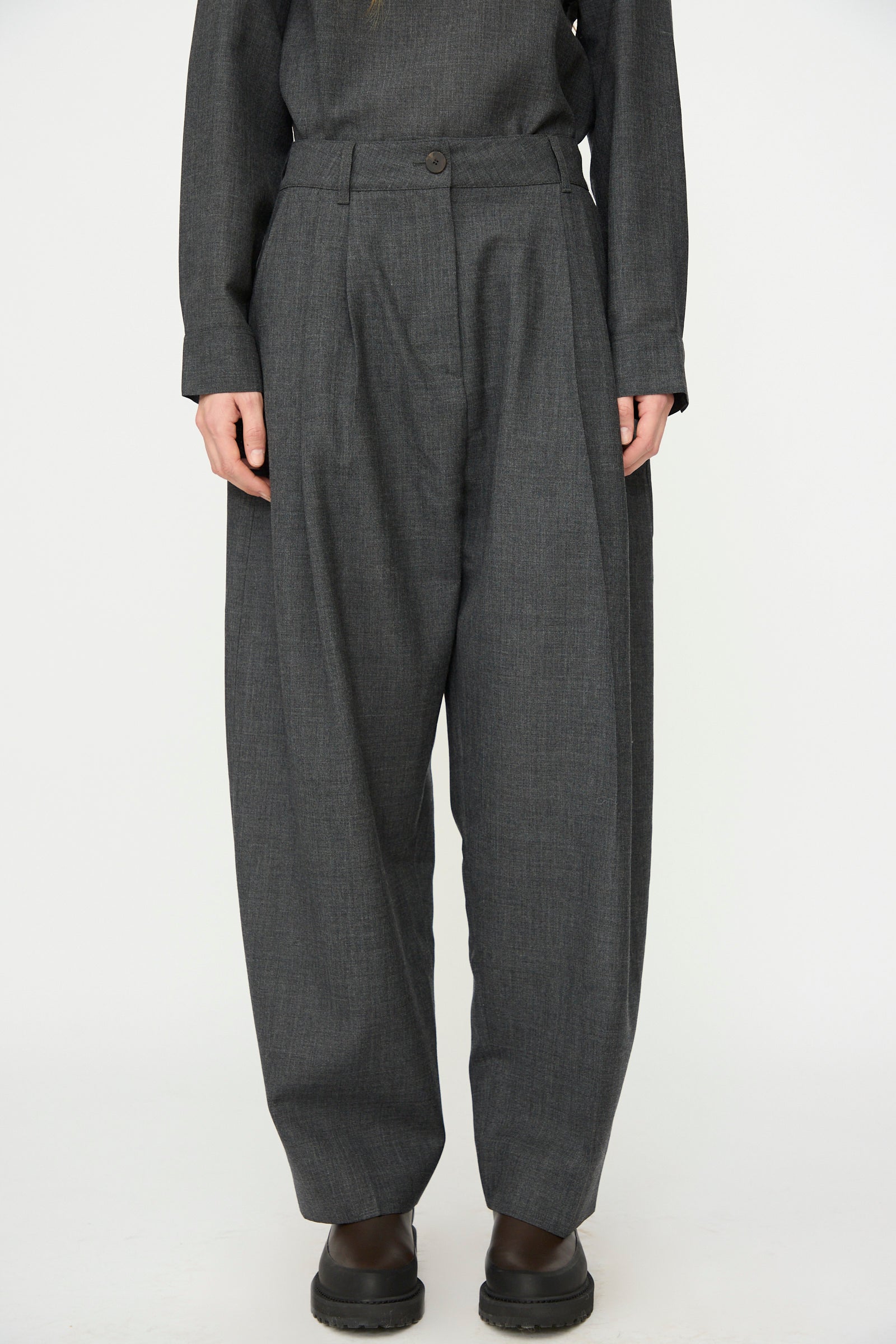 A person wearing Studio Nicholson's Heritage Wool Acuna Pant in Slate Melange, paired with a matching long-sleeve top and black shoes, stands against a plain white background.