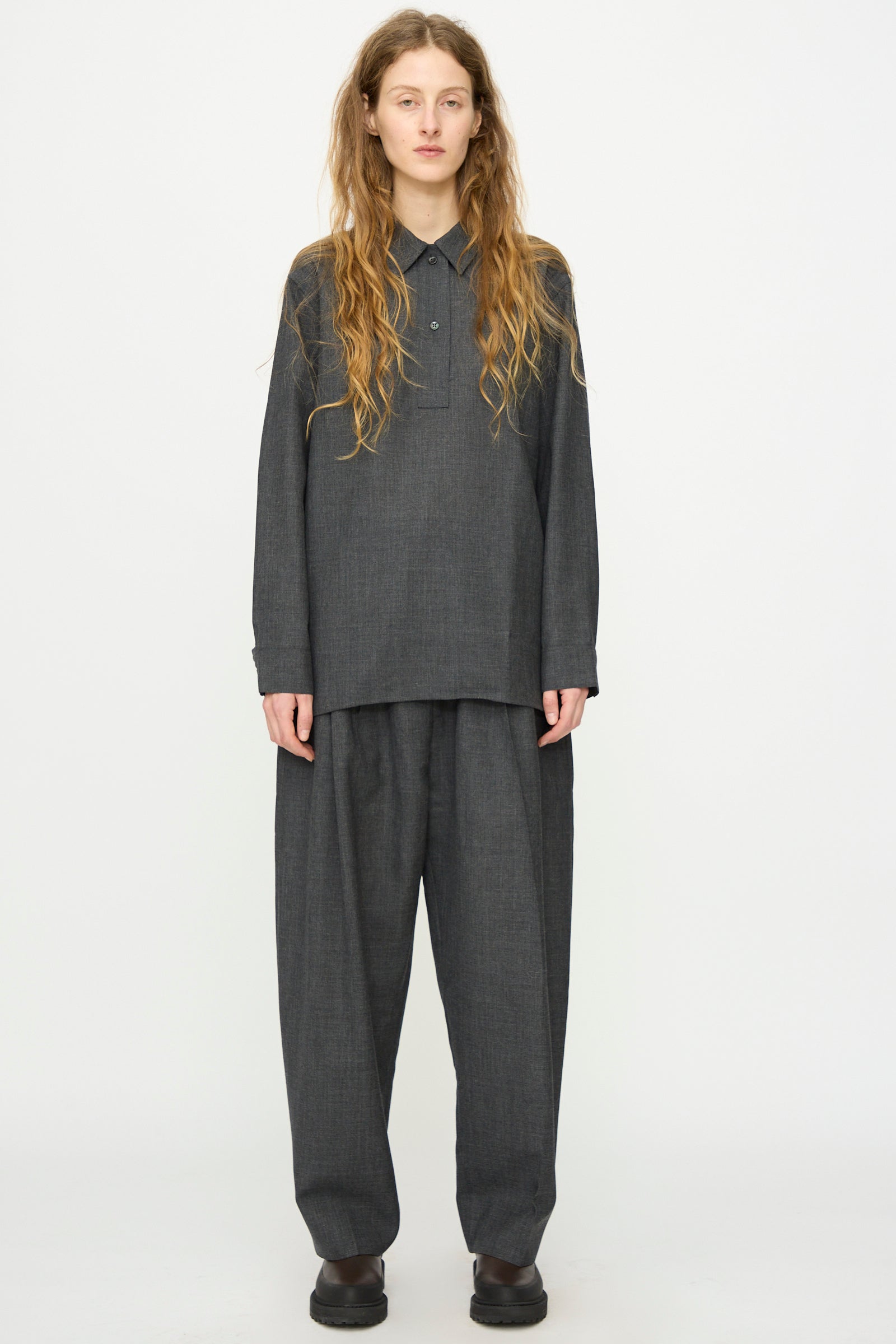 A person with long hair wears the Studio Nicholson Heritage Wool Pinole Shirt in Slate Melange, styled with matching wide-leg trousers, standing against a plain white background.