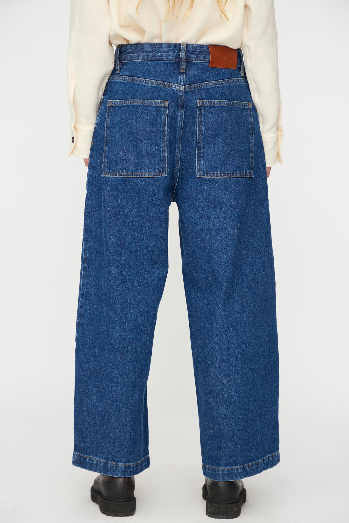 A person stands facing away in Studio Nicholson's Neive Curved Pant in Indigo Wash, featuring wide legs and large back pockets, styled with a cream long-sleeve shirt and black shoes against a plain background.