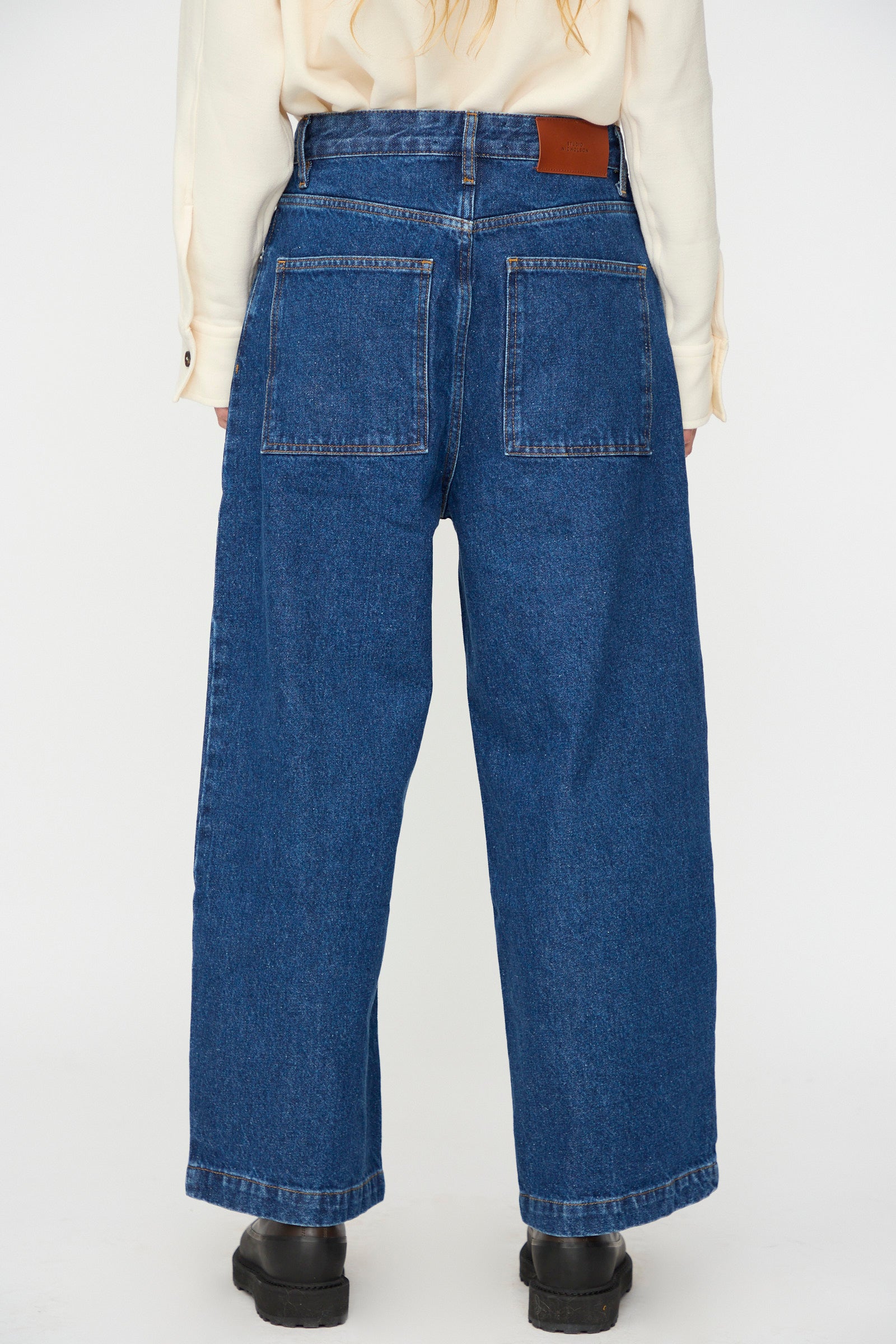 A person stands facing away in Studio Nicholson's Neive Curved Pant in Indigo Wash, featuring wide legs and large back pockets, styled with a cream long-sleeve shirt and black shoes against a plain background.