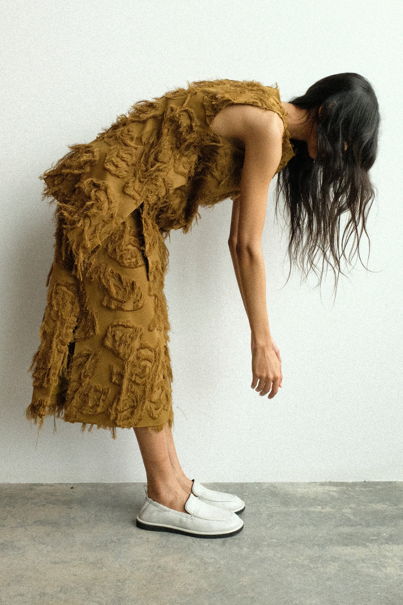A woman bending down wearing Studio Nicholson's Fringe Jacquard Mager Top in Mustard with the Fringe Jacquard Roeper Skirt in Mustard against a white wall.