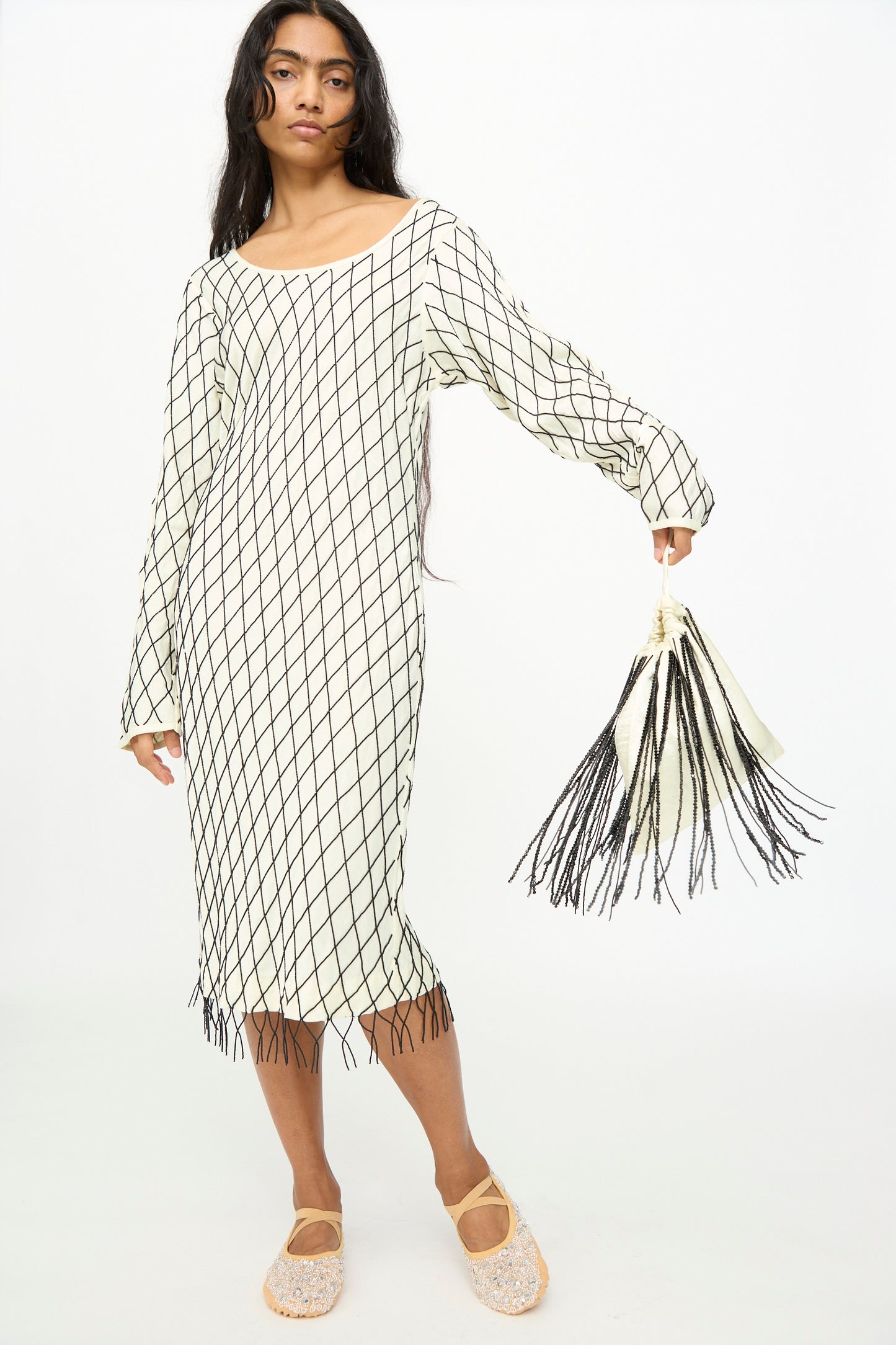A person stands wearing a long-sleeve, white dress with a black grid pattern and fringe, beige shoes, and holds a matching accessory—a TIGRA TIGRA Beaded Fringe Wristlet Bag in pale yellow with black beads, handmade in India.