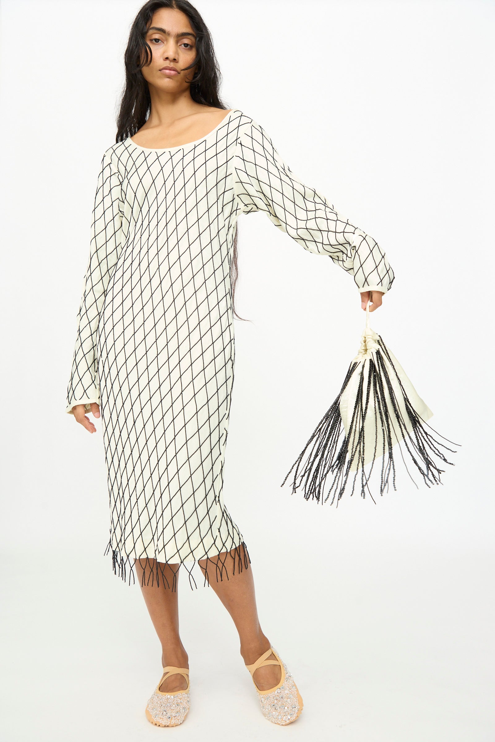 A person stands wearing a long-sleeve, white dress with a black grid pattern and fringe, beige shoes, and holds a matching accessory—a TIGRA TIGRA Beaded Fringe Wristlet Bag in pale yellow with black beads, handmade in India.