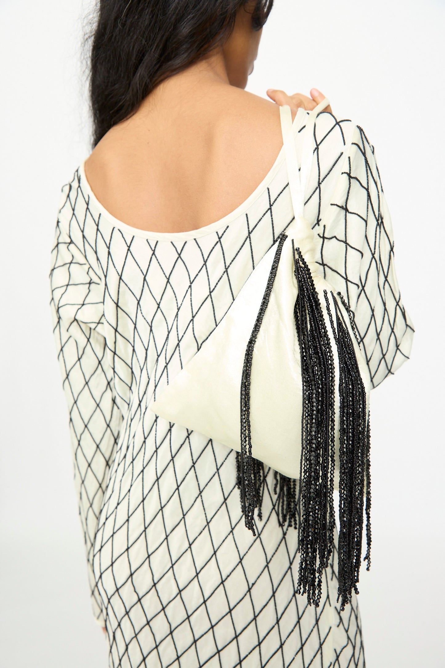 A woman with long dark hair, wearing a white dress with a black grid pattern, carries the TIGRA TIGRA Beaded Fringe Wristlet Bag in pale yellow, handmade in India and adorned with black beaded fringe.